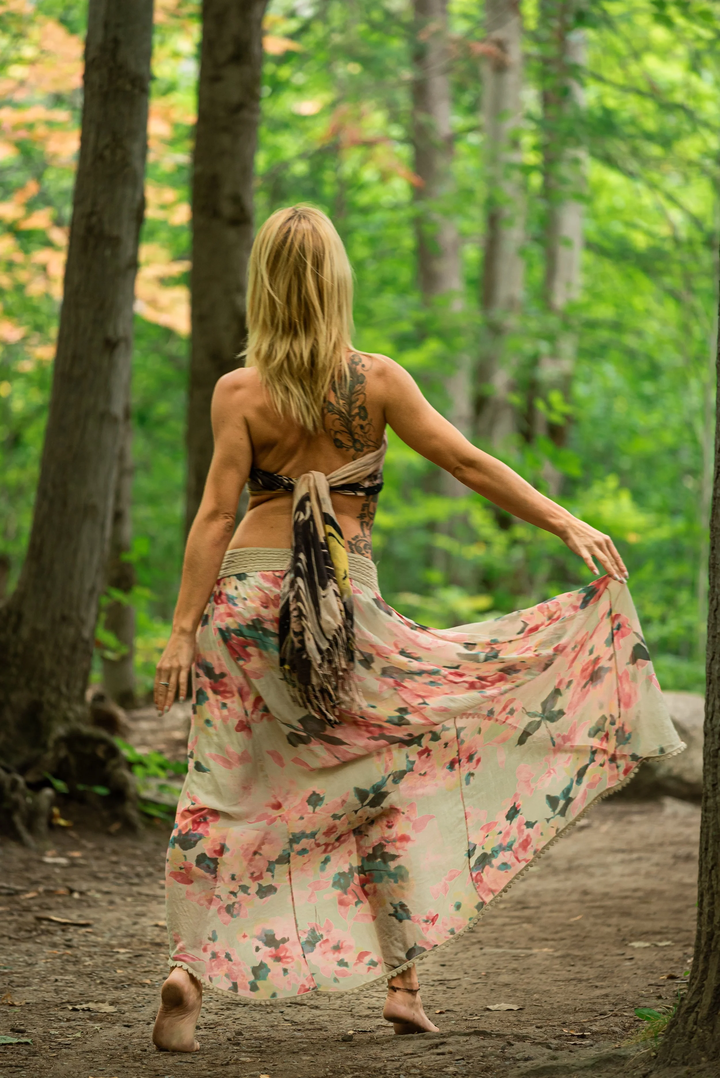 A woman with blonde hair and tattoos is walking barefoot on a dirt path through a dense, green forest, holding up her floral skirt.
