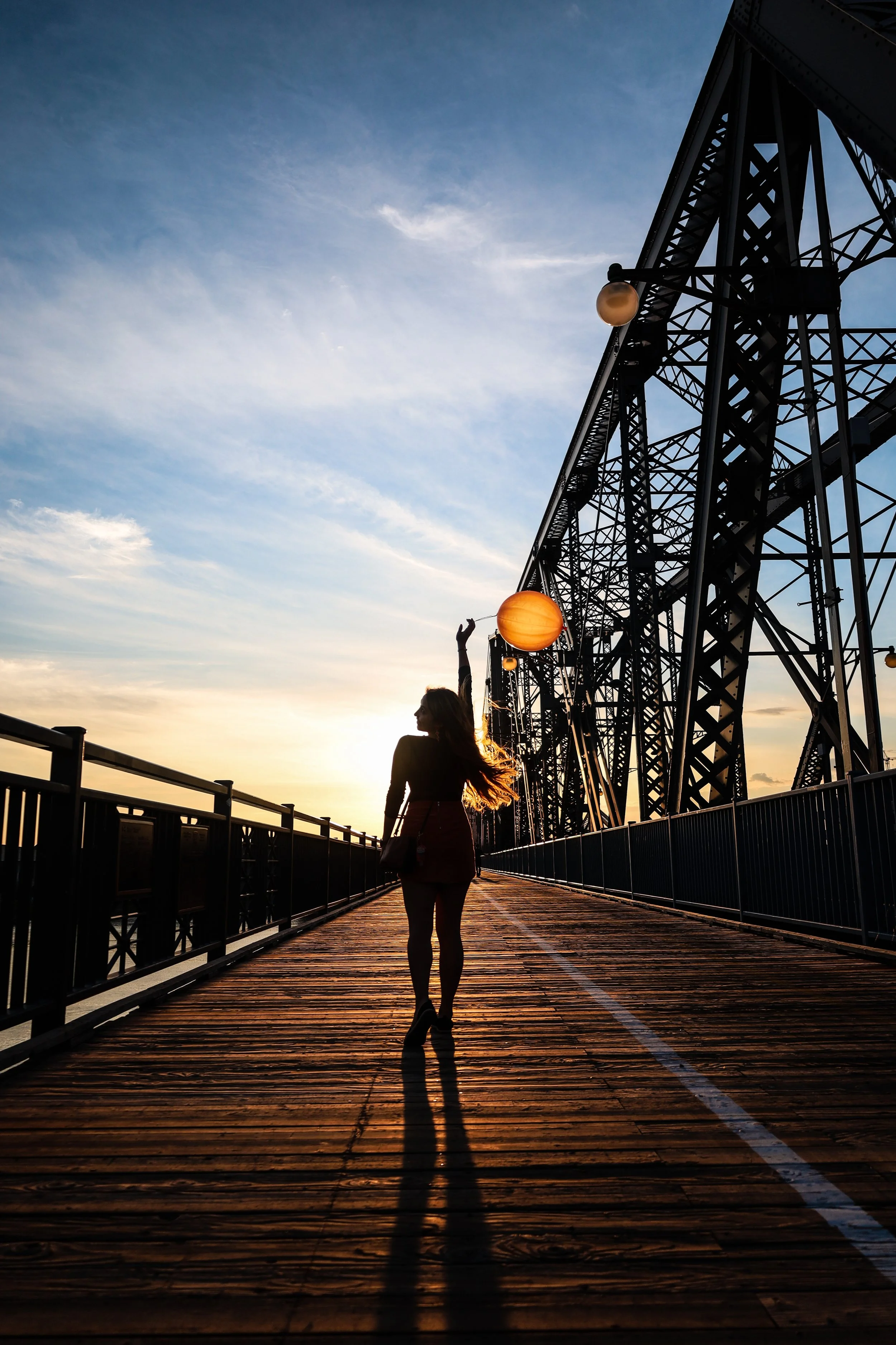 Silhouette of a woman walking on a wooden bridge at sunset, with an industrial bridge structure and hanging lamps in the background.