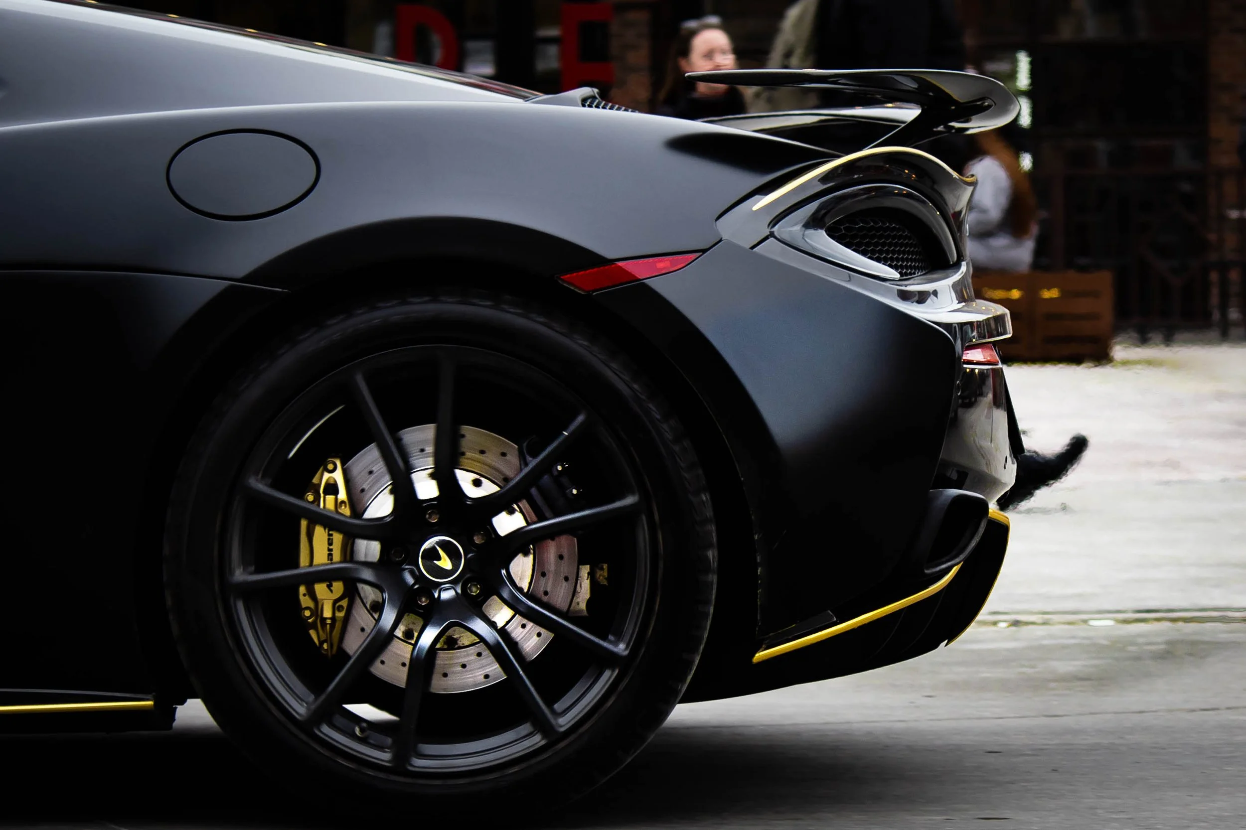 Close-up of a black sports car with yellow accents, showing the front wheel and part of the front bumper, in a parking area.