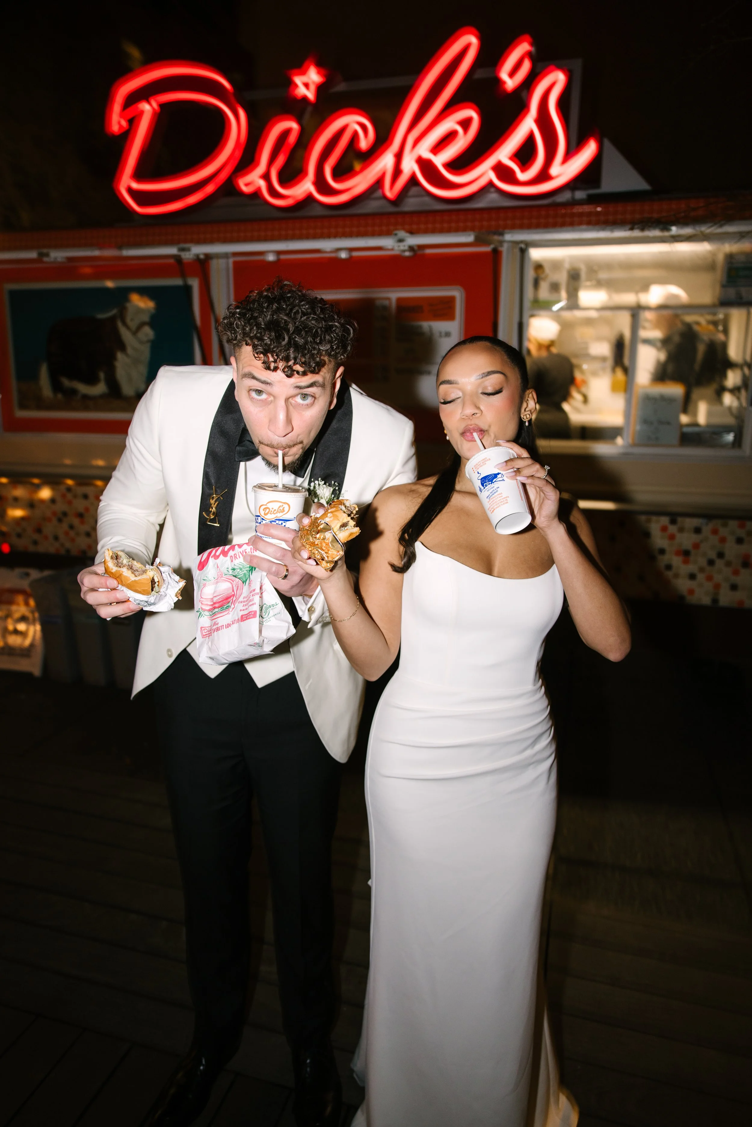 A man and woman in formal wedding attire enjoying burgers and milkshake in the foreground with the Dick's Drive-In's food truck in the background at night, with a neon rooftop sign that says 'Dick's' glowing on top.