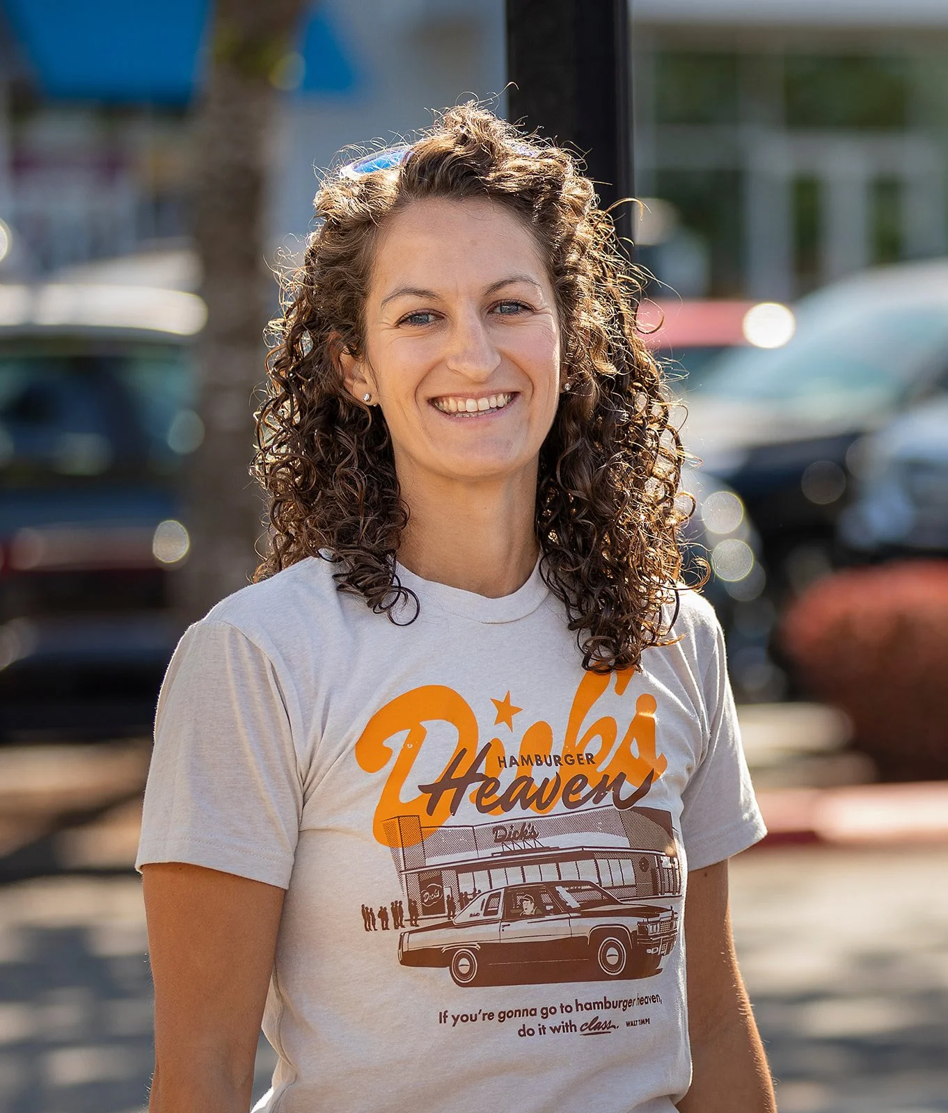 Jasmine Donovan, Dick's Drive-In President & CEO, smiling outdoors and wearing the vintage-style "Hamburger Heaven" T-shirt, featuring a classic car in front of the Crossroads Drive-In location.