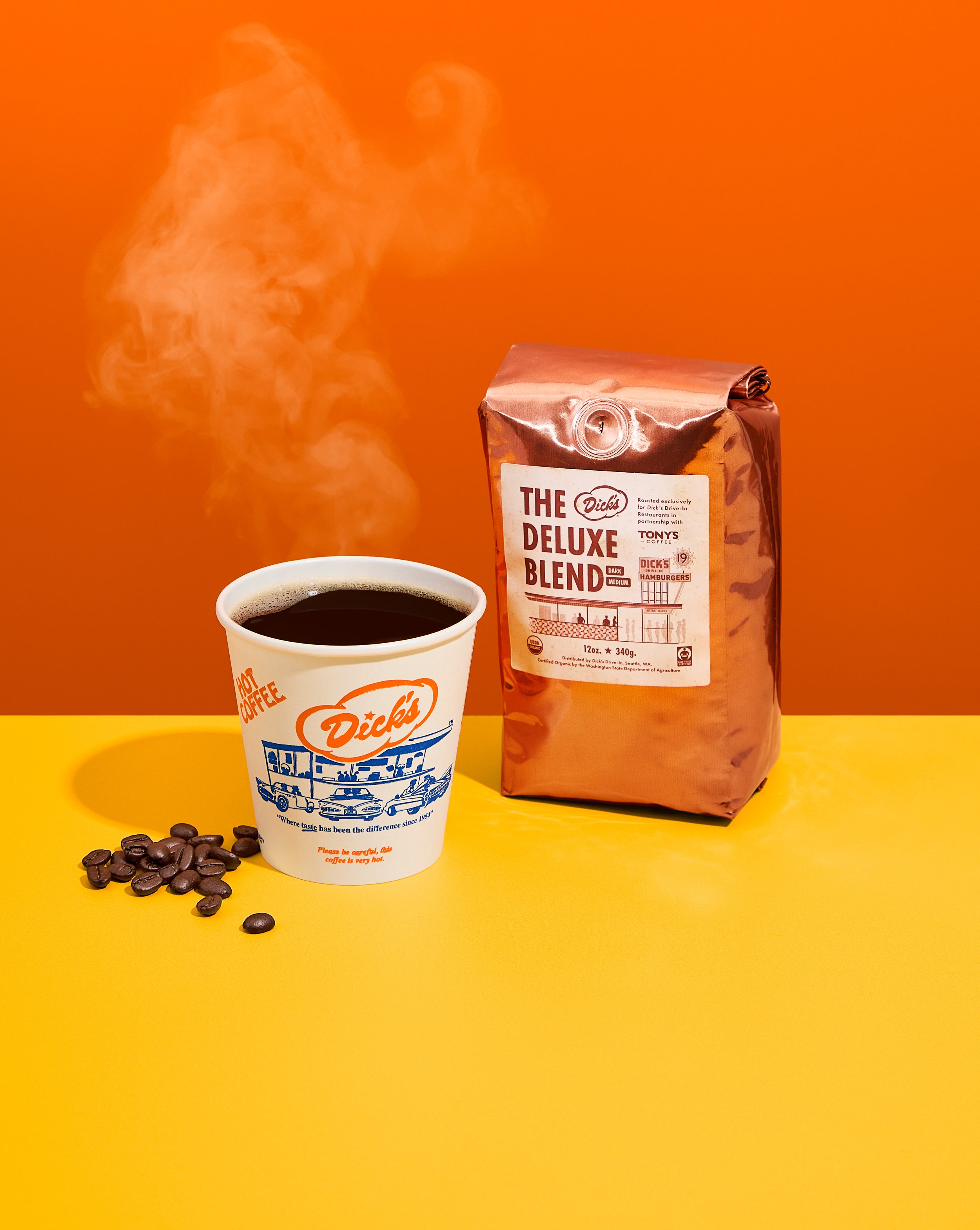 A Dick's Drive-In cup of black coffee with rising steam. Next to it is a bag of Dick's Deluxe Blend with some coffee beans scattered near the cup, all placed on a vibrant yellow surface with orange background.