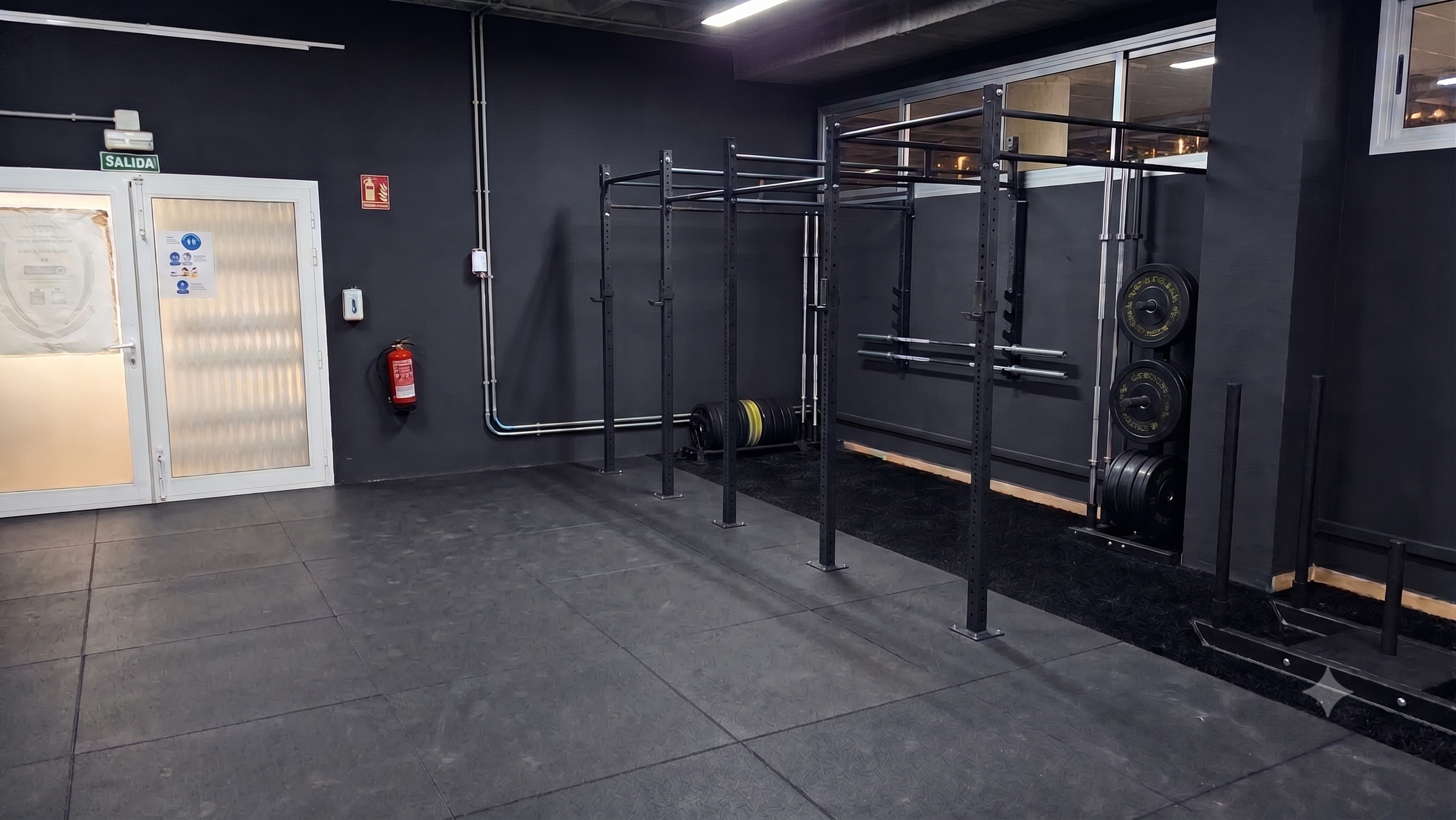 Área de gimnasio con estructura para entrenamiento de fuerza, pesas y barra, en un espacio con paredes oscuras y piso de baldosas grises.
