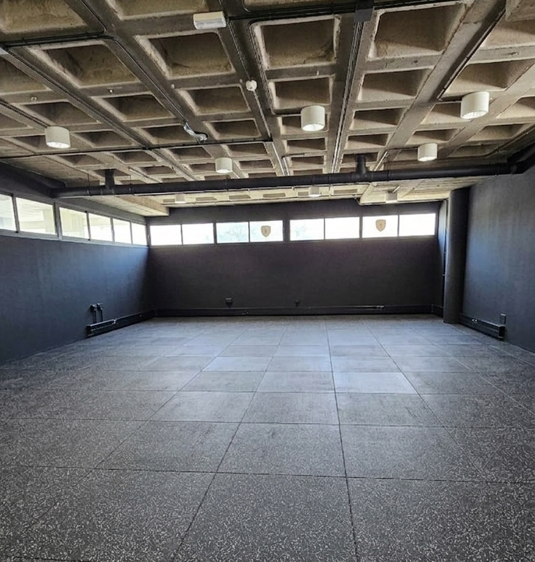 Sala vacía con paredes grises, ventanas grandes, techo con baremos y luces, suelo de baldosas y paredes laterales azules.