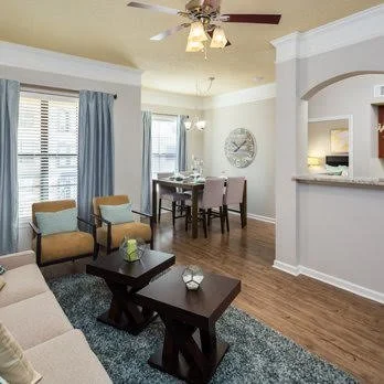 Living room and dining area with windows, blue curtains, beige walls, hardwood floor, ceiling fan, and a watch on the wall.