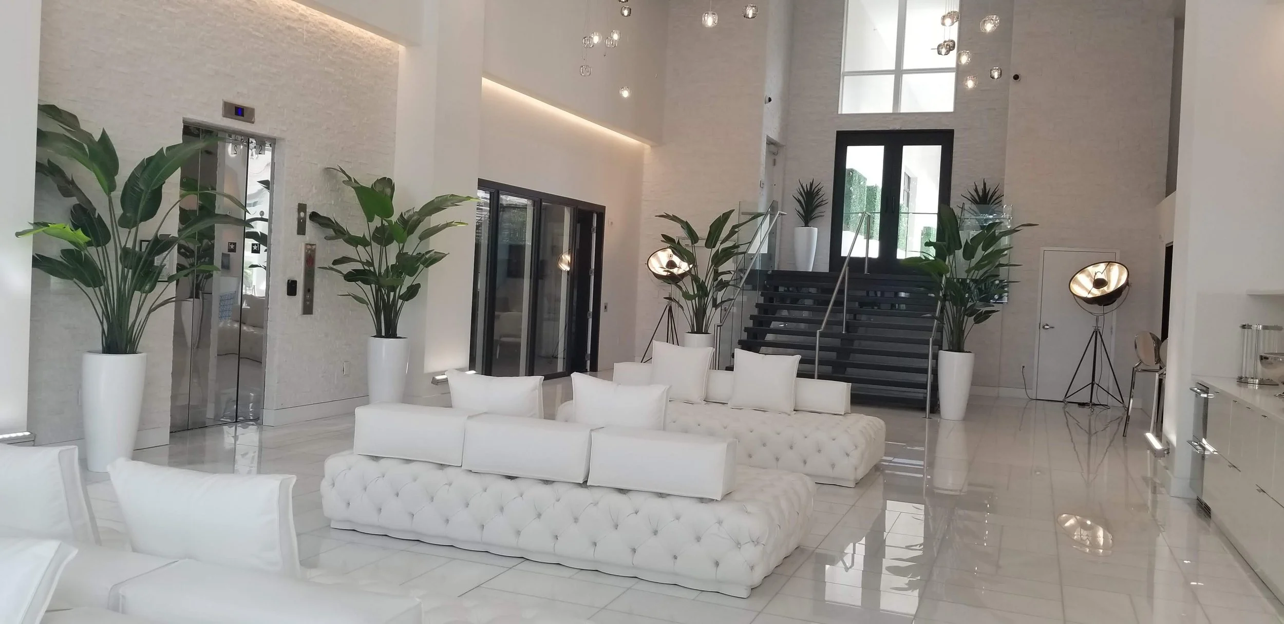 Modern hotel lobby with white tufted sofa, white chairs, large green potted plants, elevators, staircase, large window, and illuminated lighting fixtures.