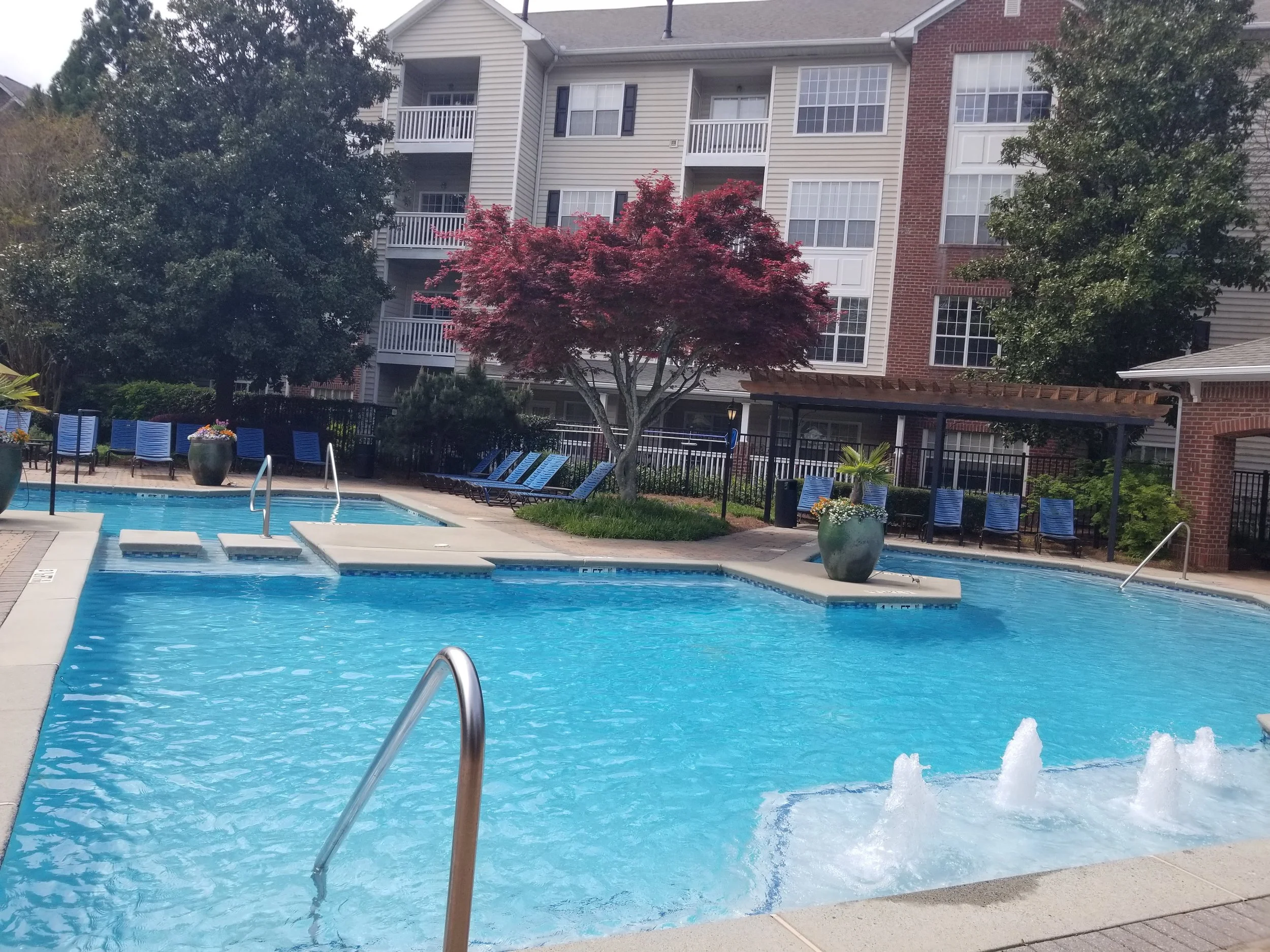 An outdoor swimming pool area with clear blue water, surrounded by lounge chairs, potted plants, and trees, including a red-leafed tree, with an apartment building in the background.