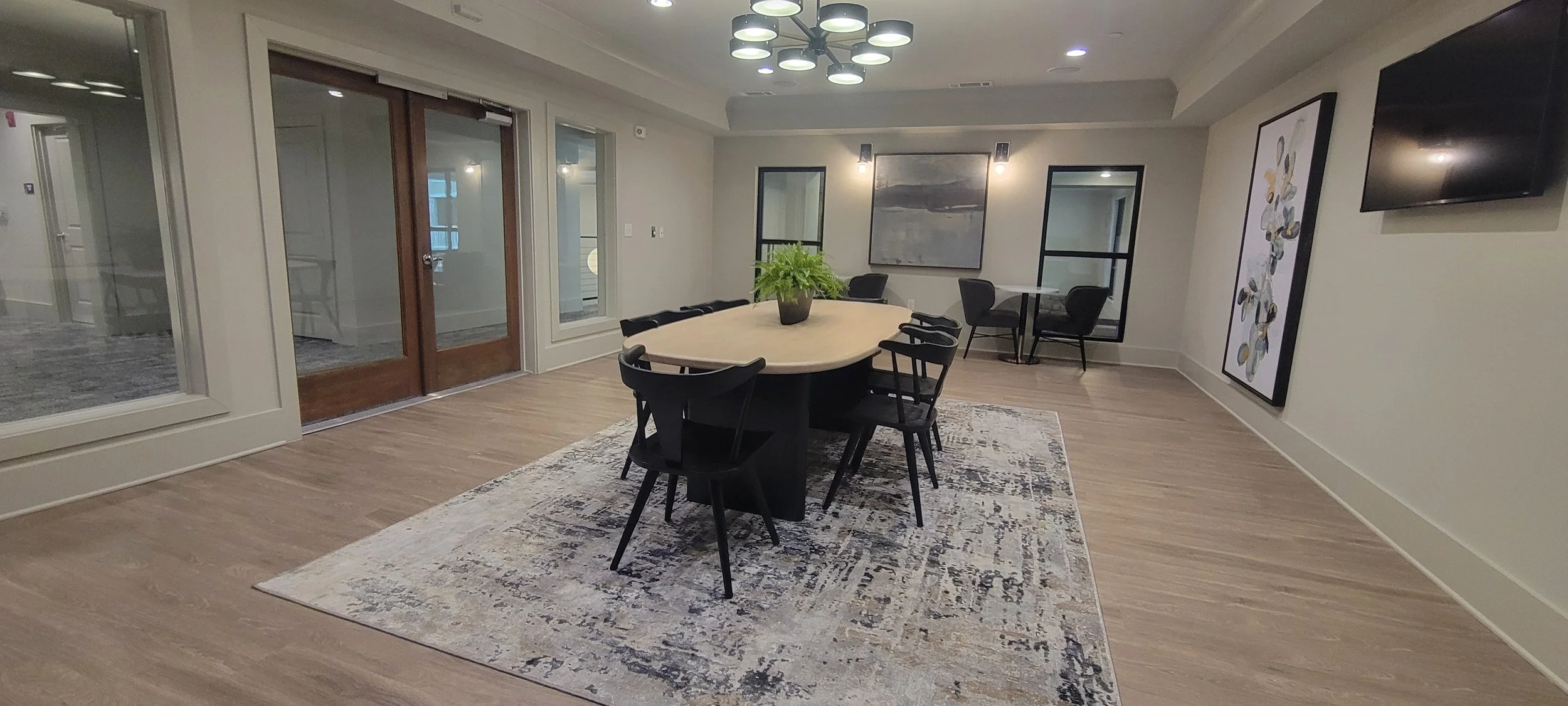Conference room with wooden table, black chairs, potted plant, artwork, large windows, and a TV on the wall.
