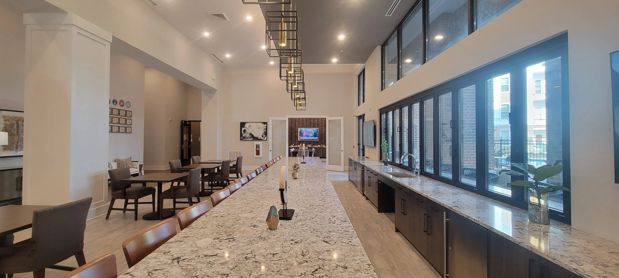 Modern communal lounge with large marble-top counter, high chairs, and a kitchen area, featuring a long row of windows and minimalistic lighting.