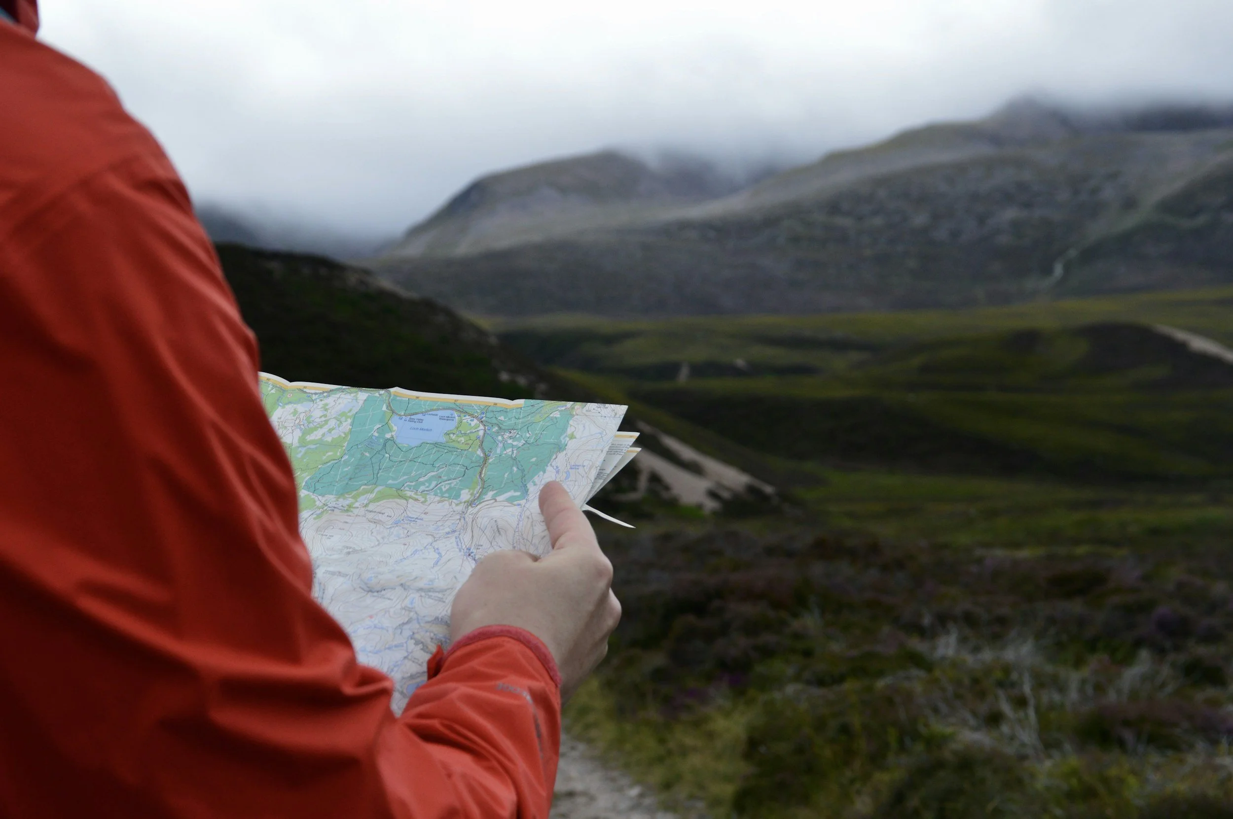 Person in red jacket holding a map while standing outdoors in a mountainous region with cloudy skies.