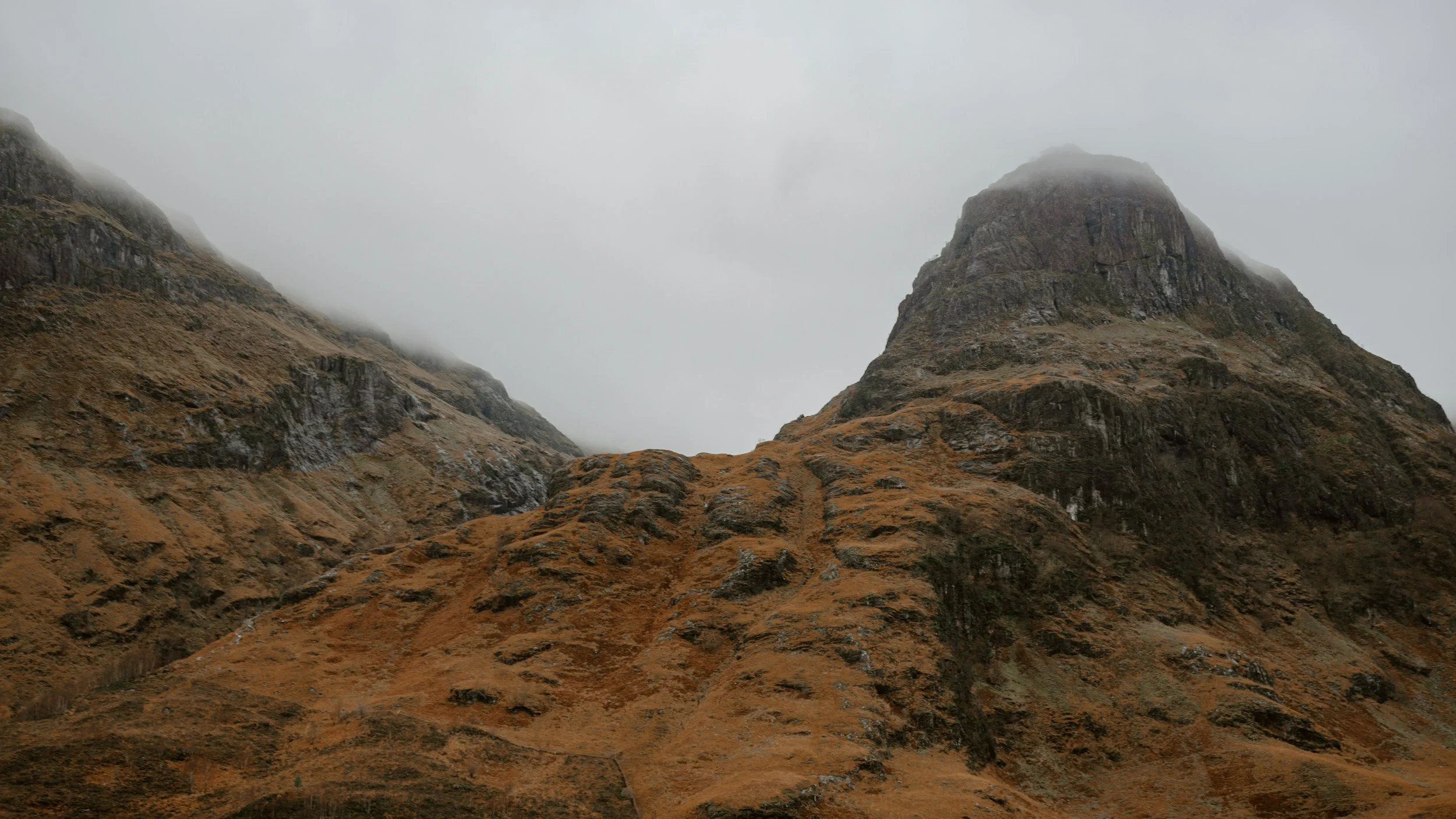 A mountainous landscape with rugged, brownish-orange slopes and steep rocky peaks. The peaks are partially obscured by low hanging clouds or fog.