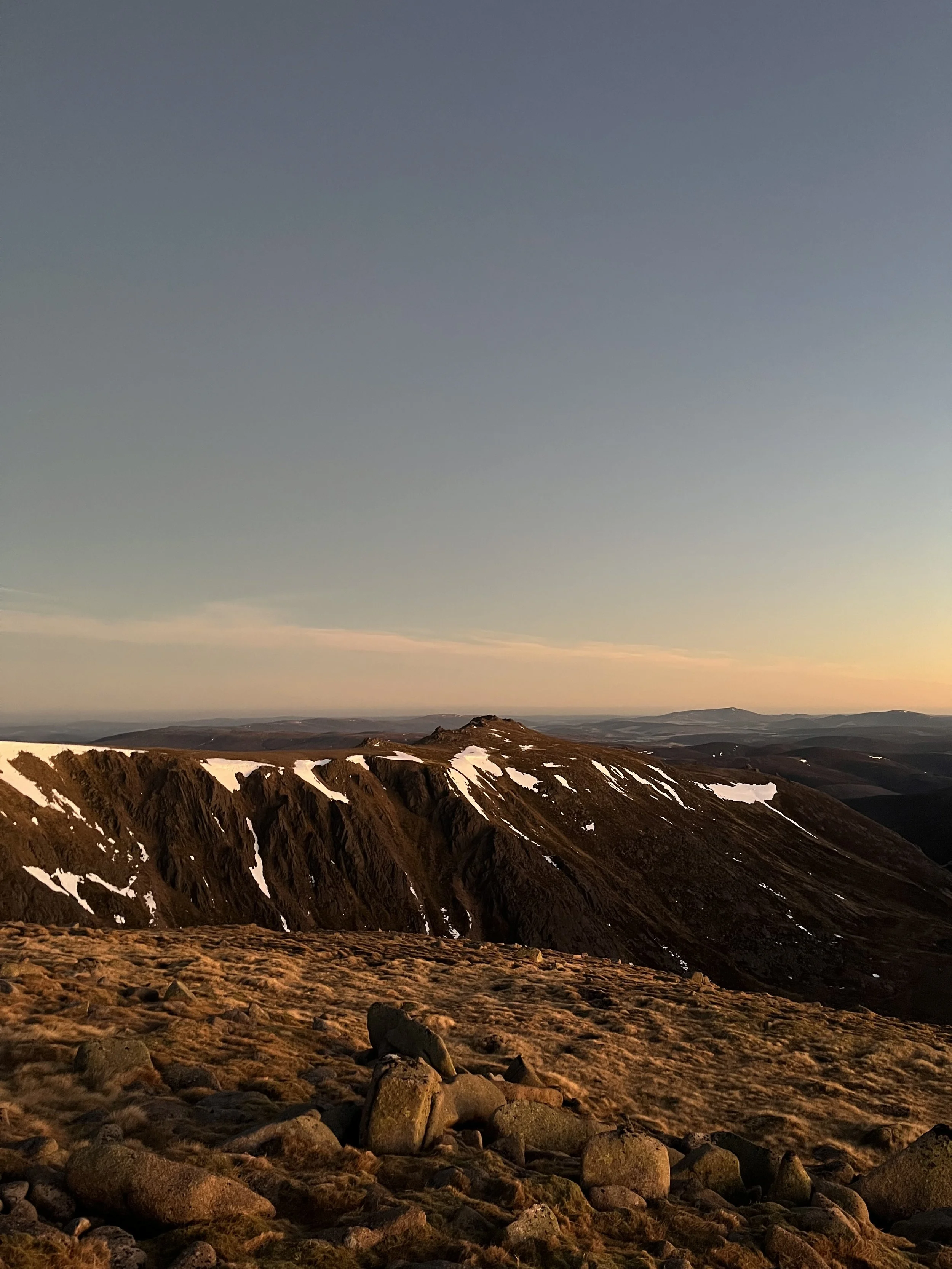 Scenic mountain landscape during sunset with rocky foreground, snow patches on mountain slopes, and a clear sky.
