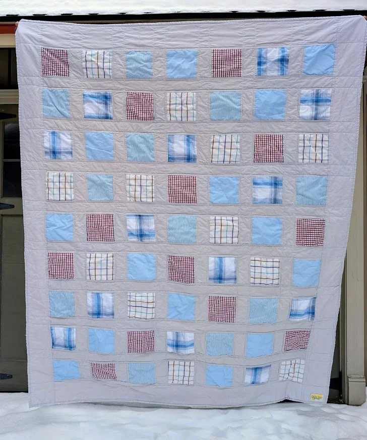 A quilt with a pattern of alternating light blue, red checkered, and multicolored checkered fabric squares, edged with a white border, hanging outdoors against a metallic surface.