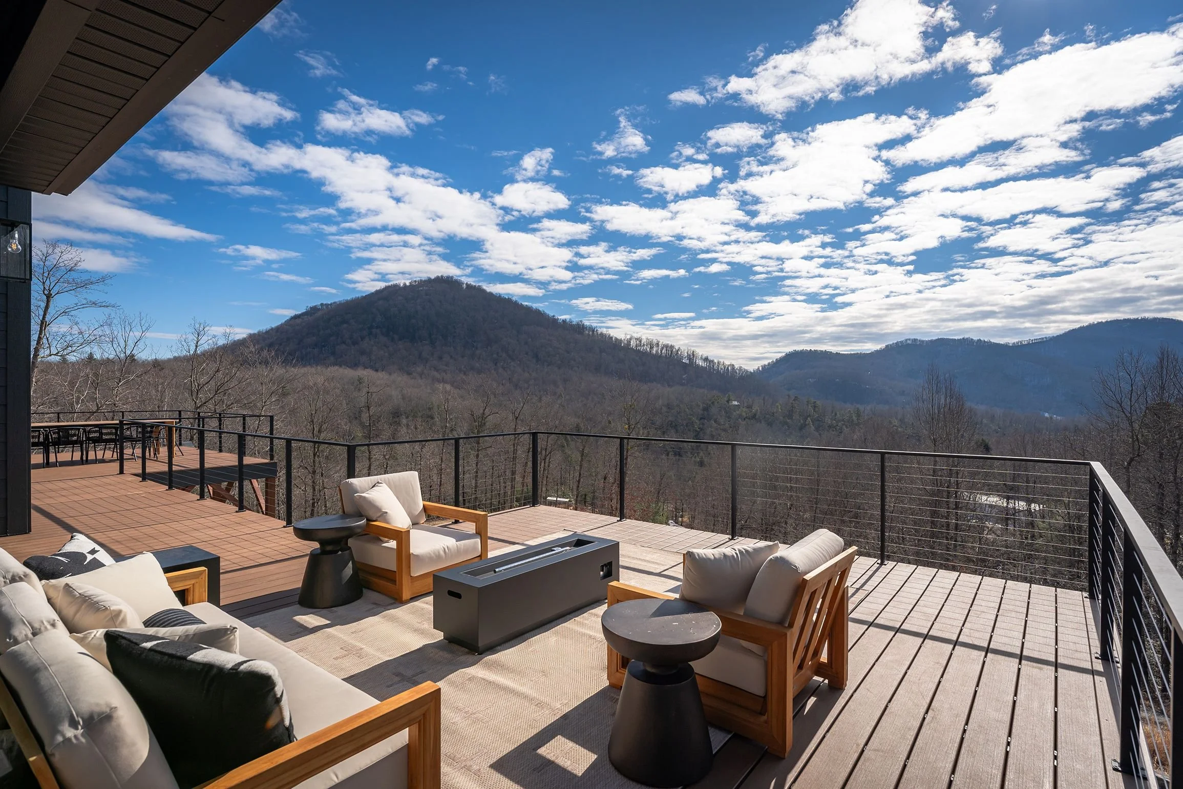 Outdoor patio with beige cushioned chairs and black side tables, overlooking a mountain landscape with trees and a partly cloudy sky.
