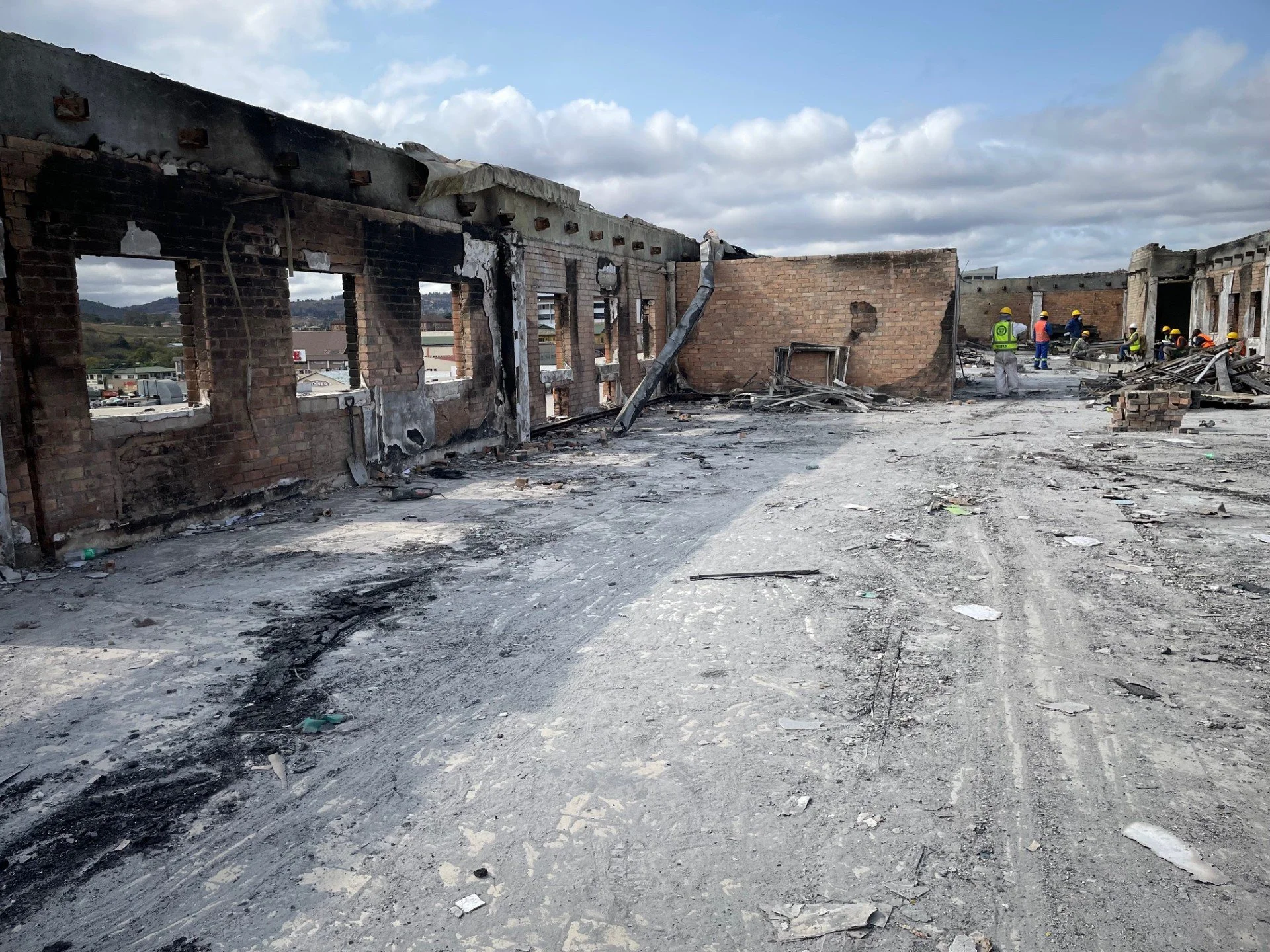 Damaged building with burnt-out walls and debris, rescue workers in yellow and orange helmets working in the background.