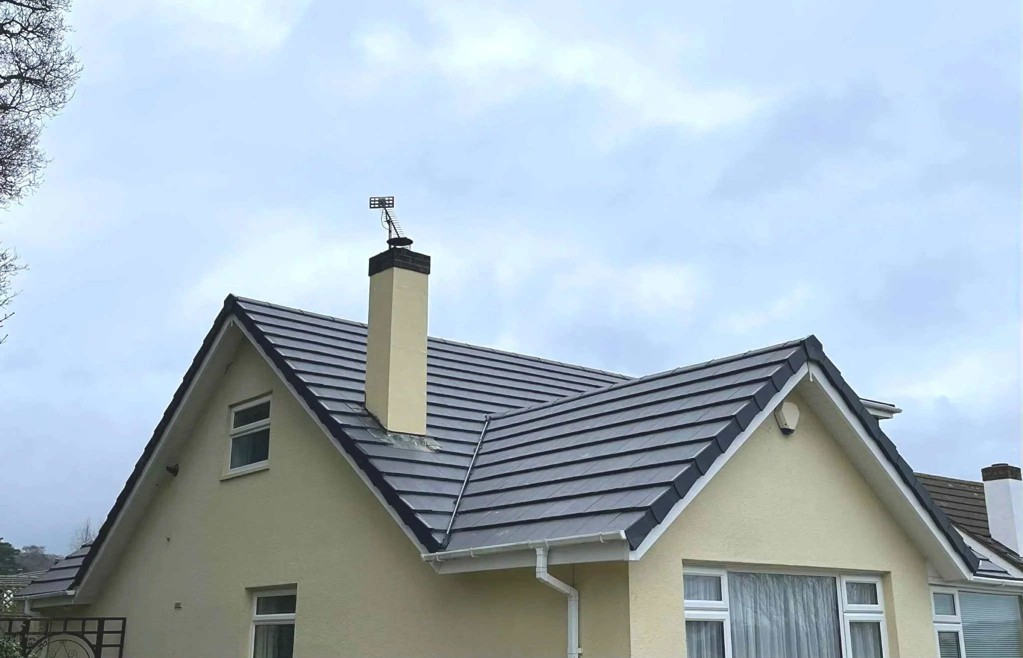 A house with light yellow exterior walls and a dark gray tiled roof. The house has a chimney with an antenna on top and a small window above. The sky is overcast with gray clouds.