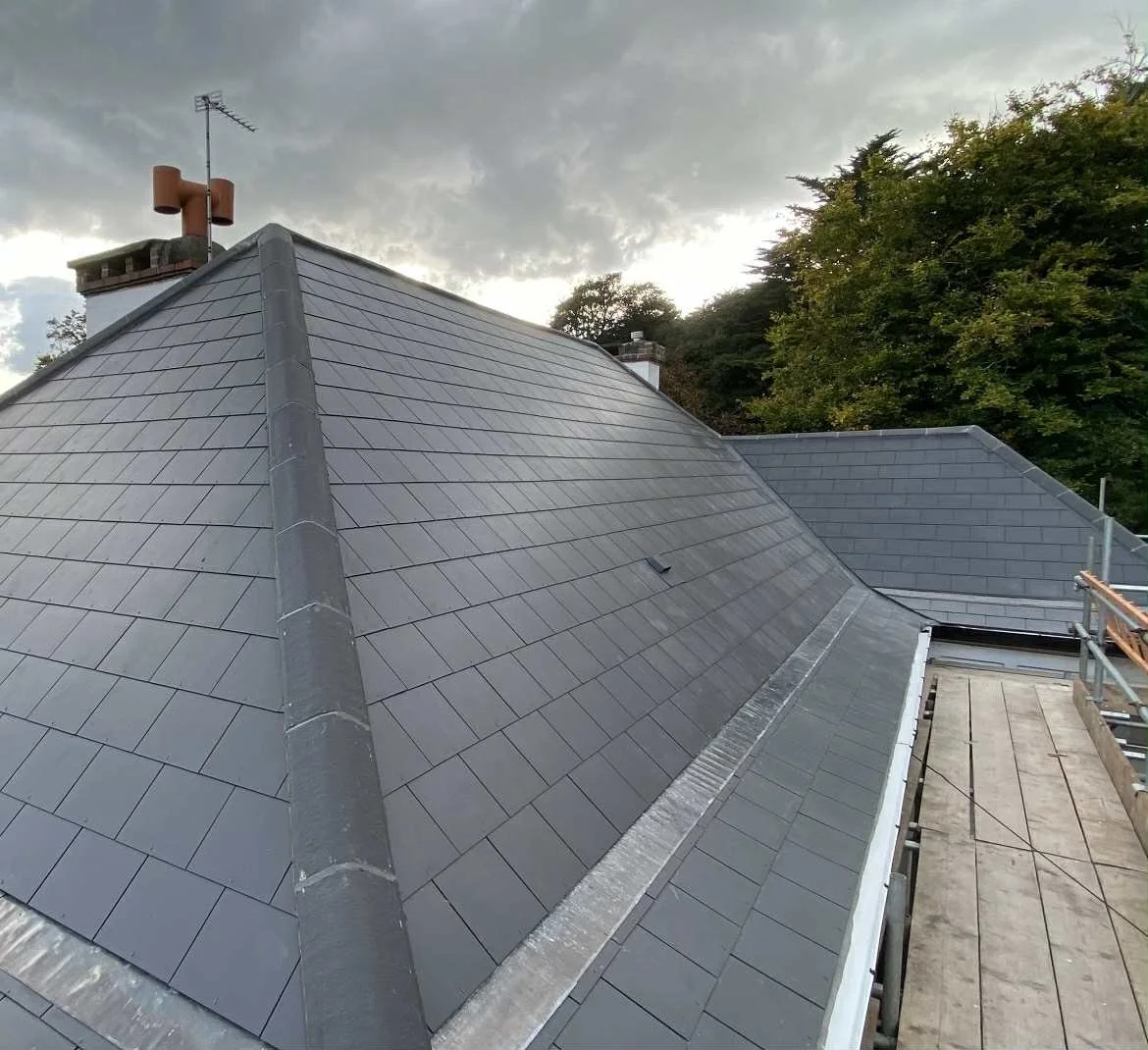 A close-up view of a grey slate tile roof on a house with a cloudy sky in the background and scaffolding on the side.