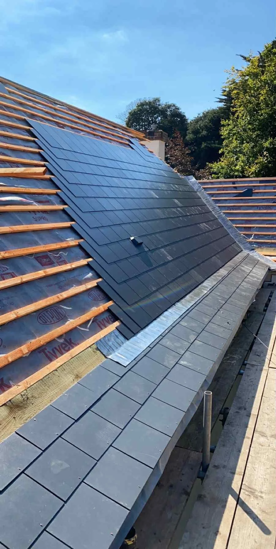 A rooftop under construction with new dark gray shingles being installed, scaffolding along the edge, and a blue sky above.