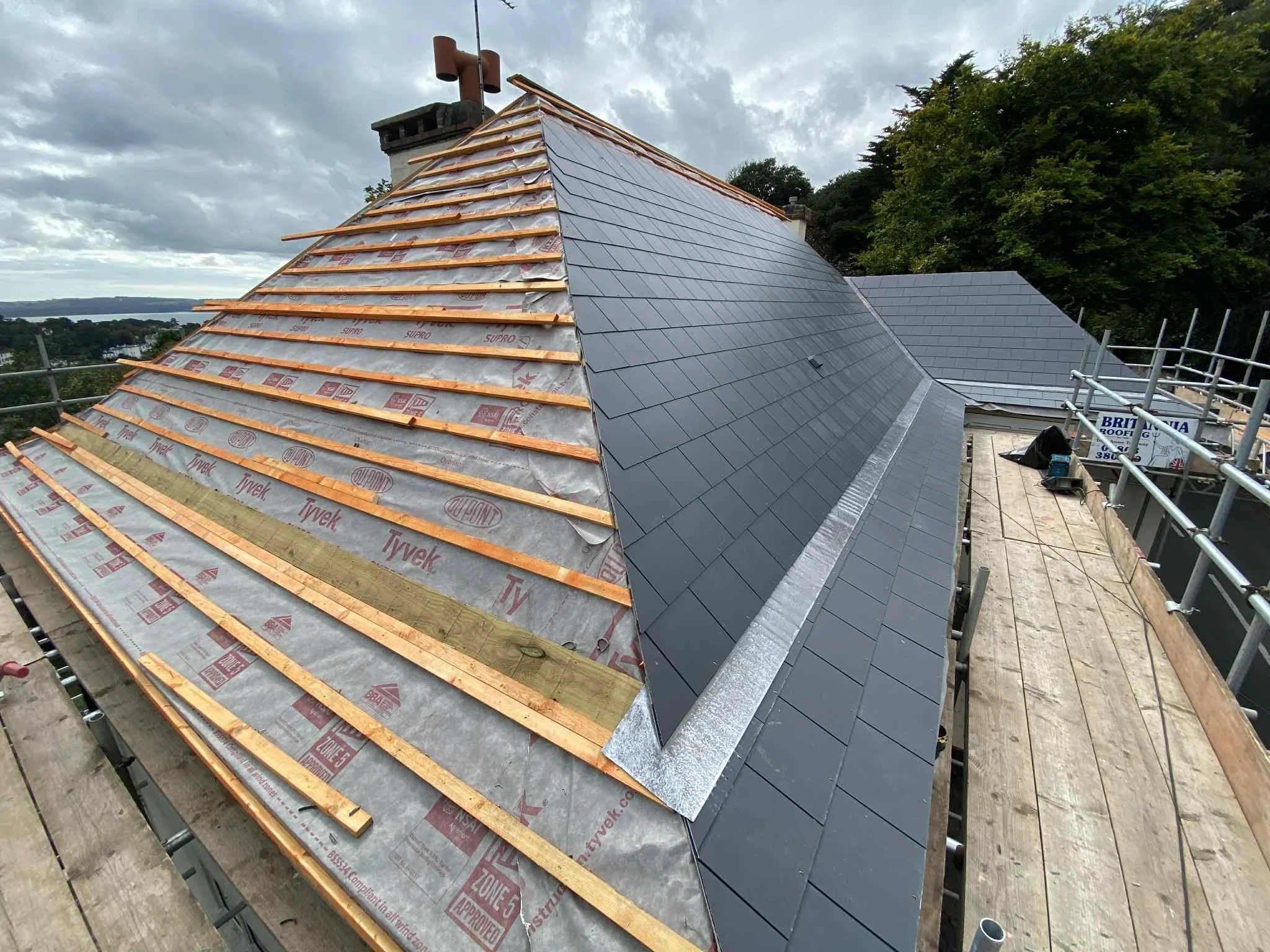 Roof under construction, with one side covered in dark tiles and the other side in progress, showing insulation and wooden battens, scaffolding around the roof, and a cloudy sky in the background.