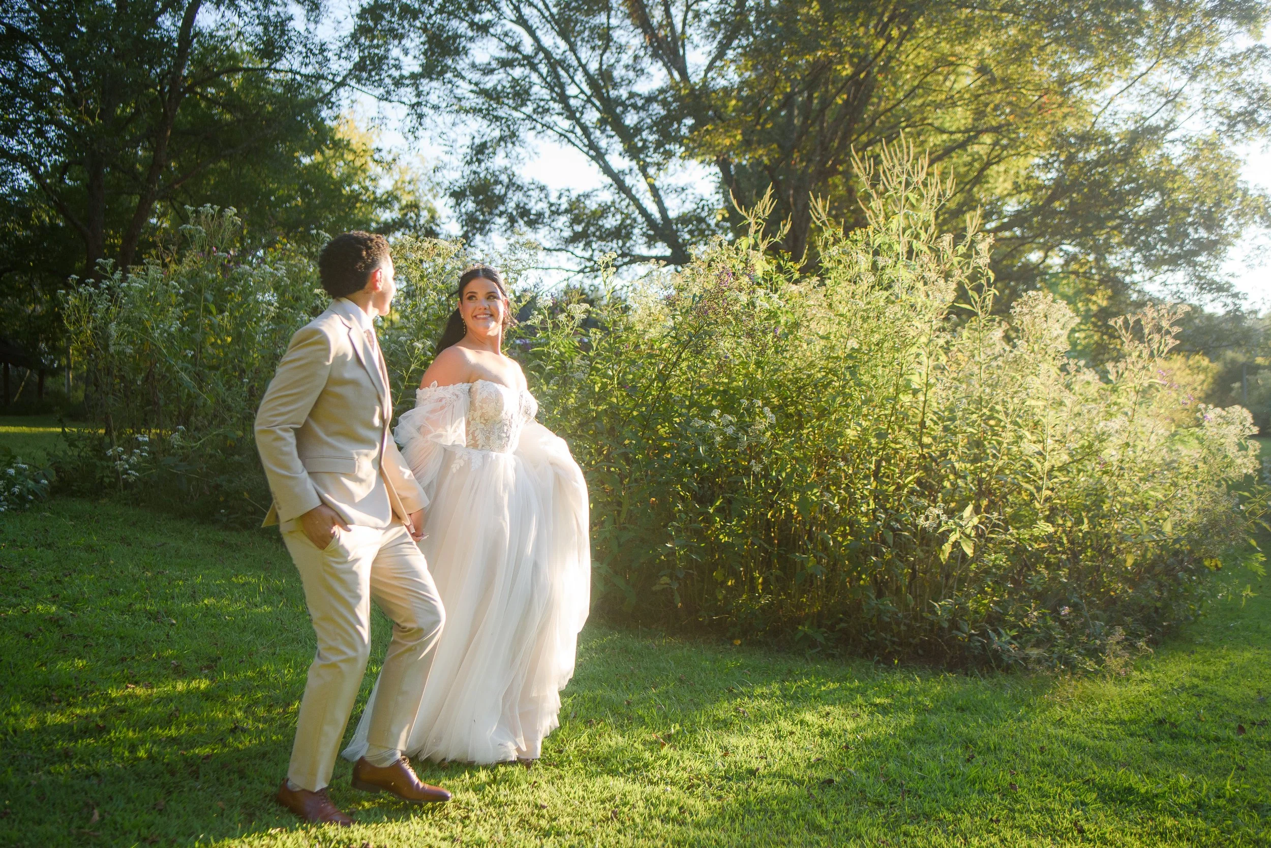 A bride and groom walk hand in hand in a lush garden during sunset, surrounded by tall trees and greenery, with the bride smiling.