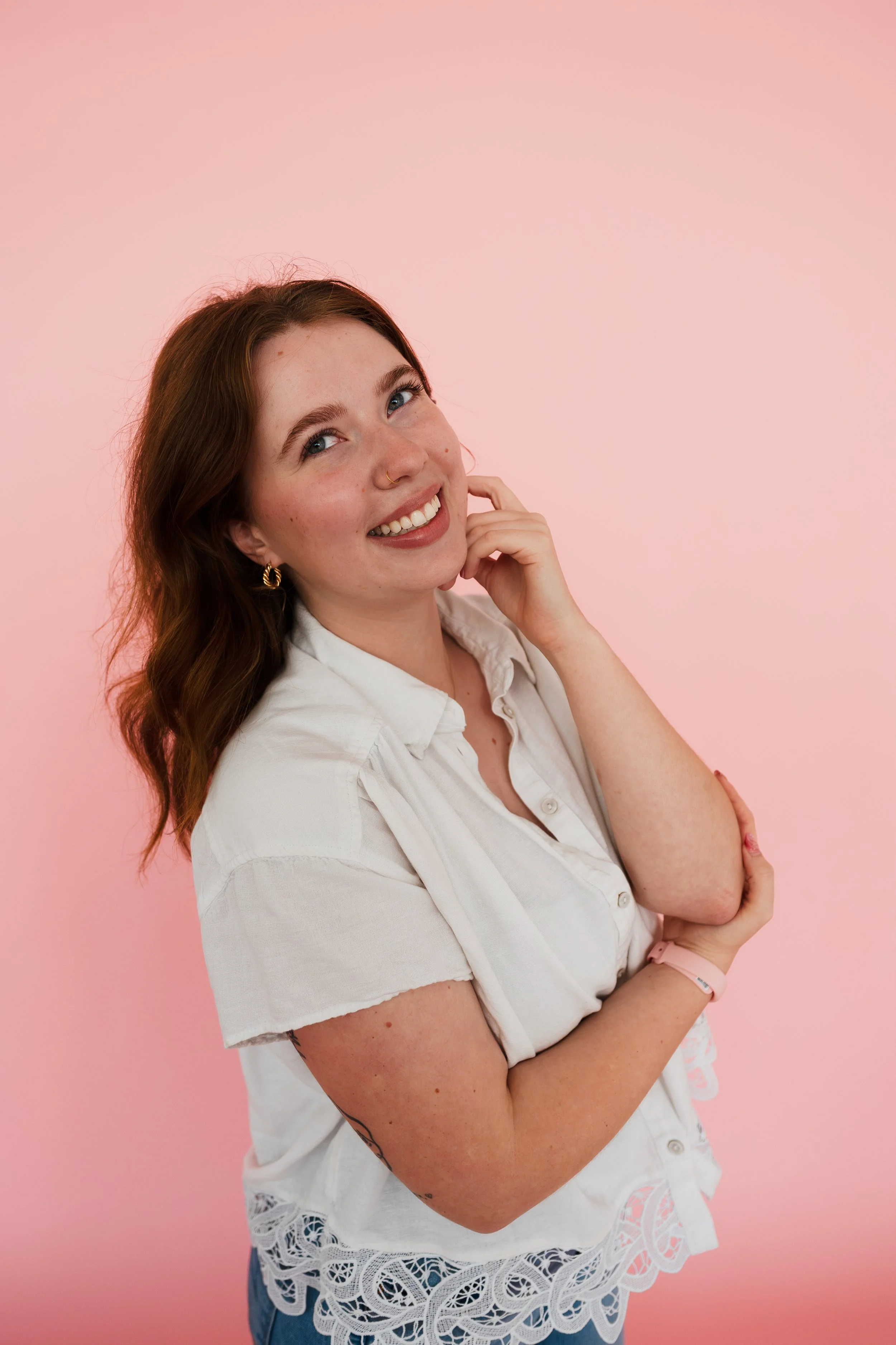 Young woman with long, wavy brown hair and blue eyes, smiling and looking slightly upward, wearing a white blouse with lace detailing and gold hoop earrings, standing against a pink background.