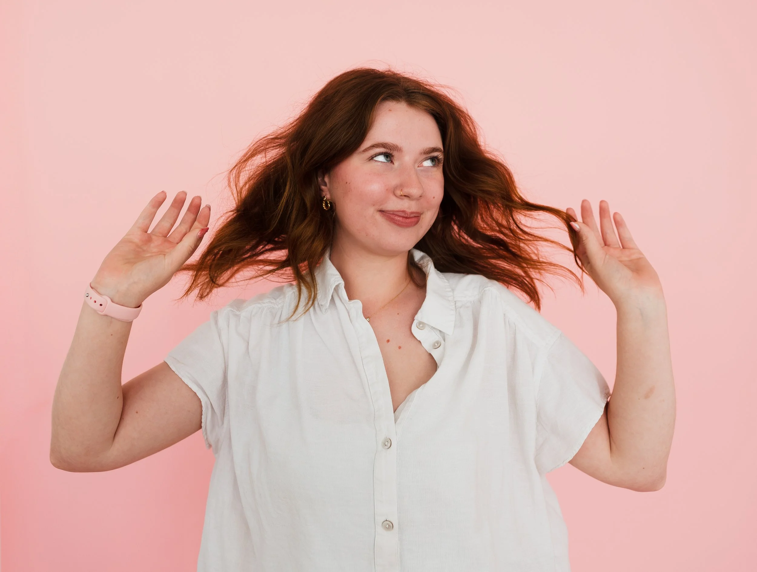 A woman with long auburn hair and piercings smiling while holding her hair, wearing a white shirt, against a pink background.