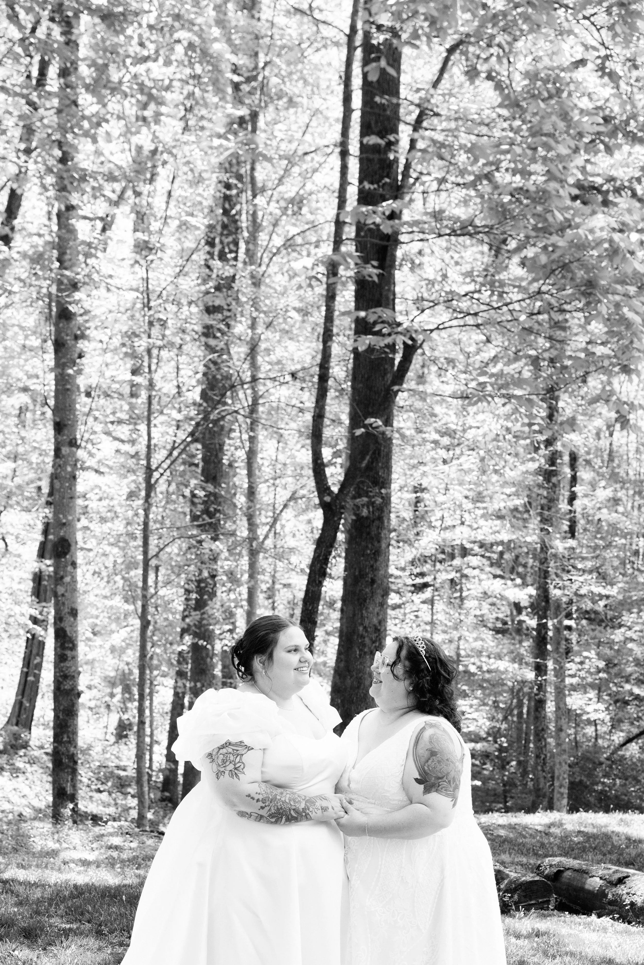 Two women in wedding dresses standing in a wooded outdoor setting, smiling and holding hands, with tall trees and foliage in the background.