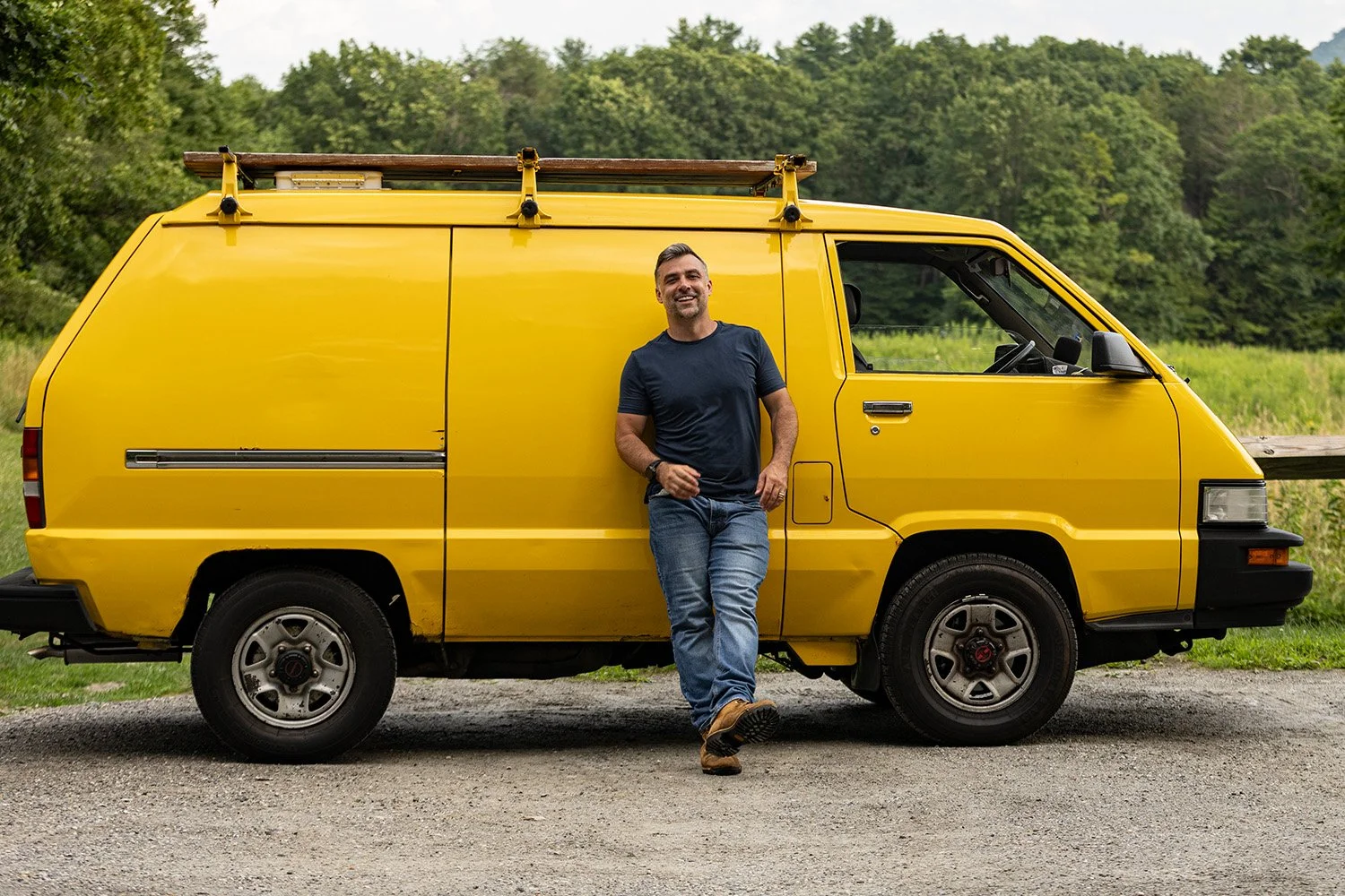 United States Congressional candidate Jeromie Whalen leaning against a yellow utility van with a ladder on top, outdoors with trees in the background.