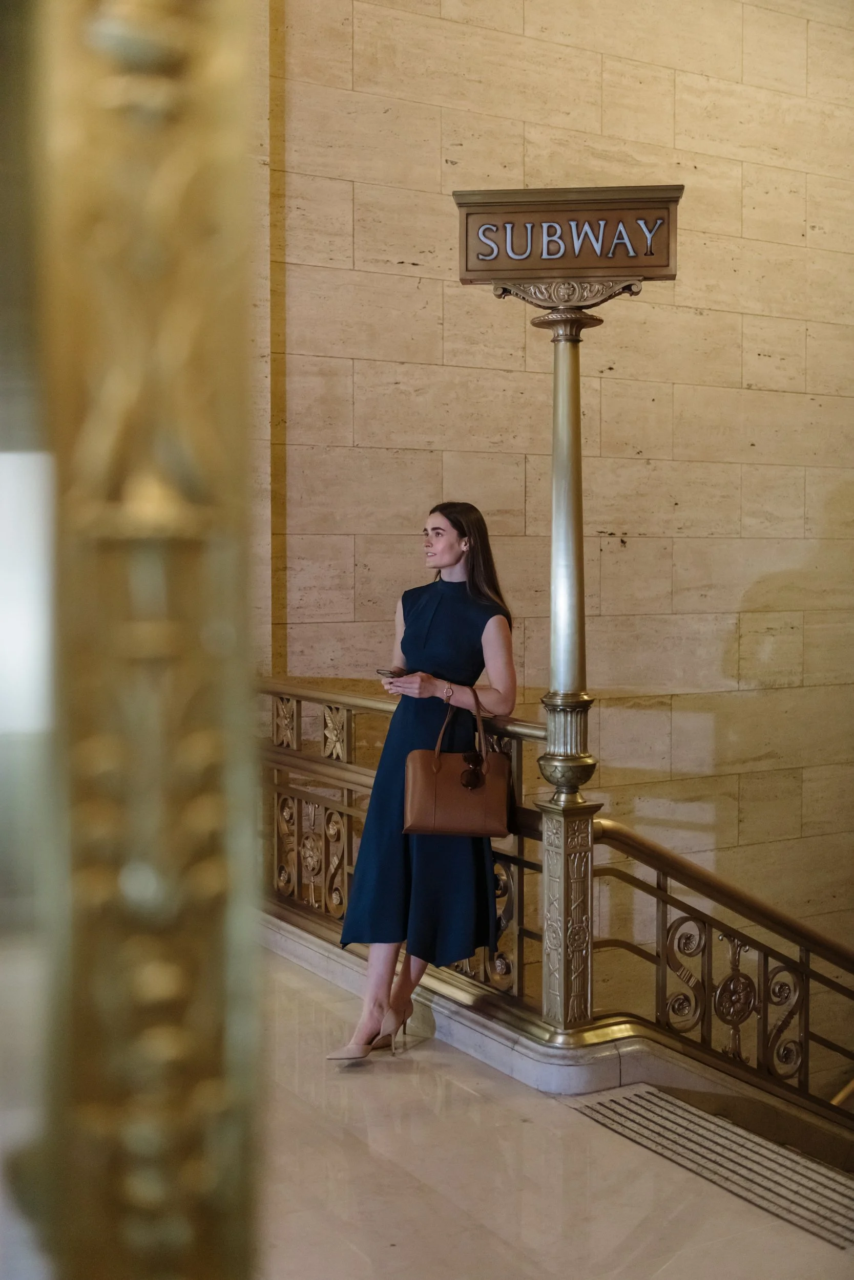 A woman is standing in front of a staircase with ornate gold railing, holding a phone and a brown handbag, next to a sign that says 'SUBWAY' in an elegant building with beige stone walls.