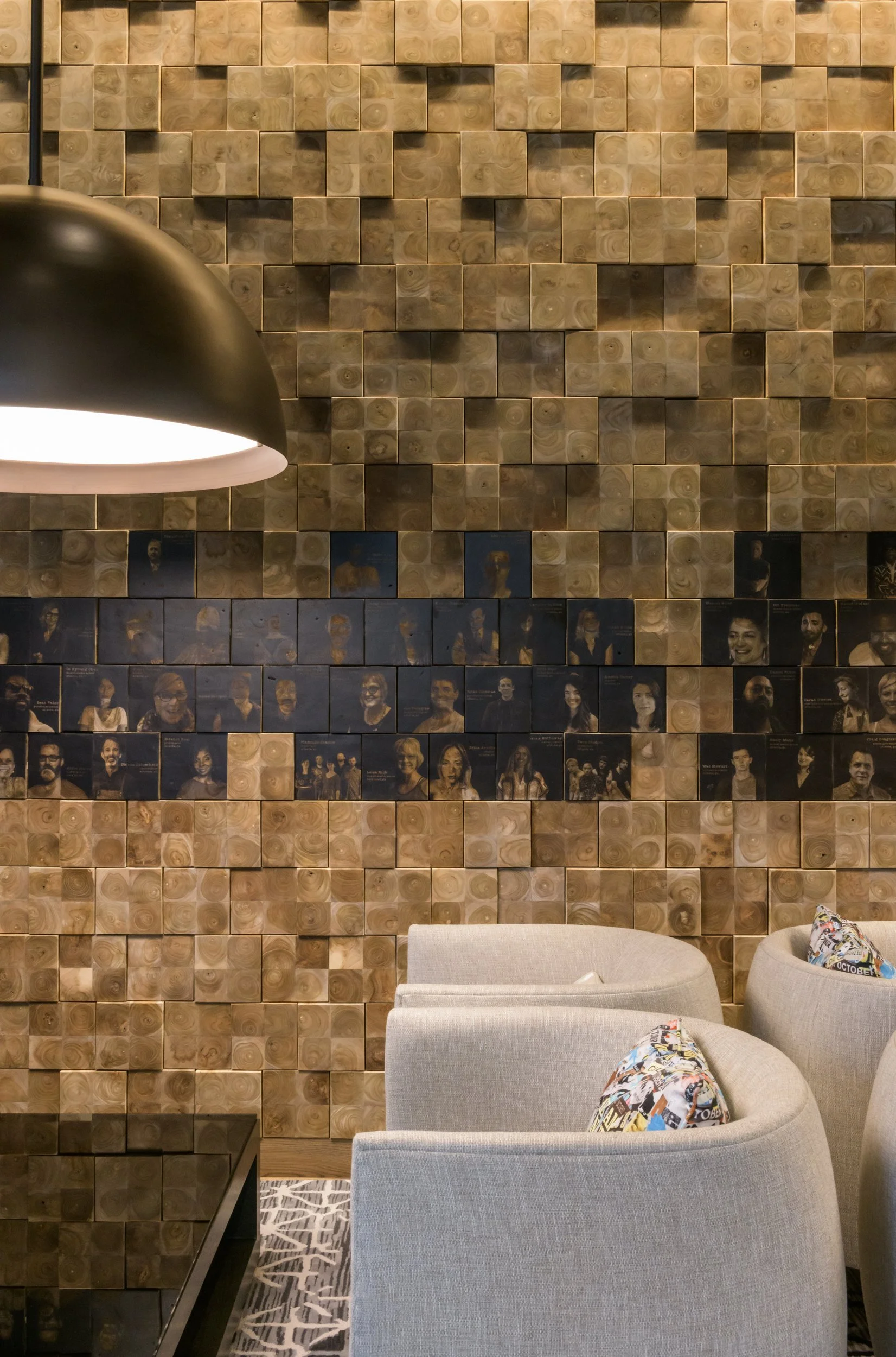 A wall decorated with small wooden cubes and blue portrait tiles of diverse individuals, with a seating area of gray armchairs and colorful pillows in front.