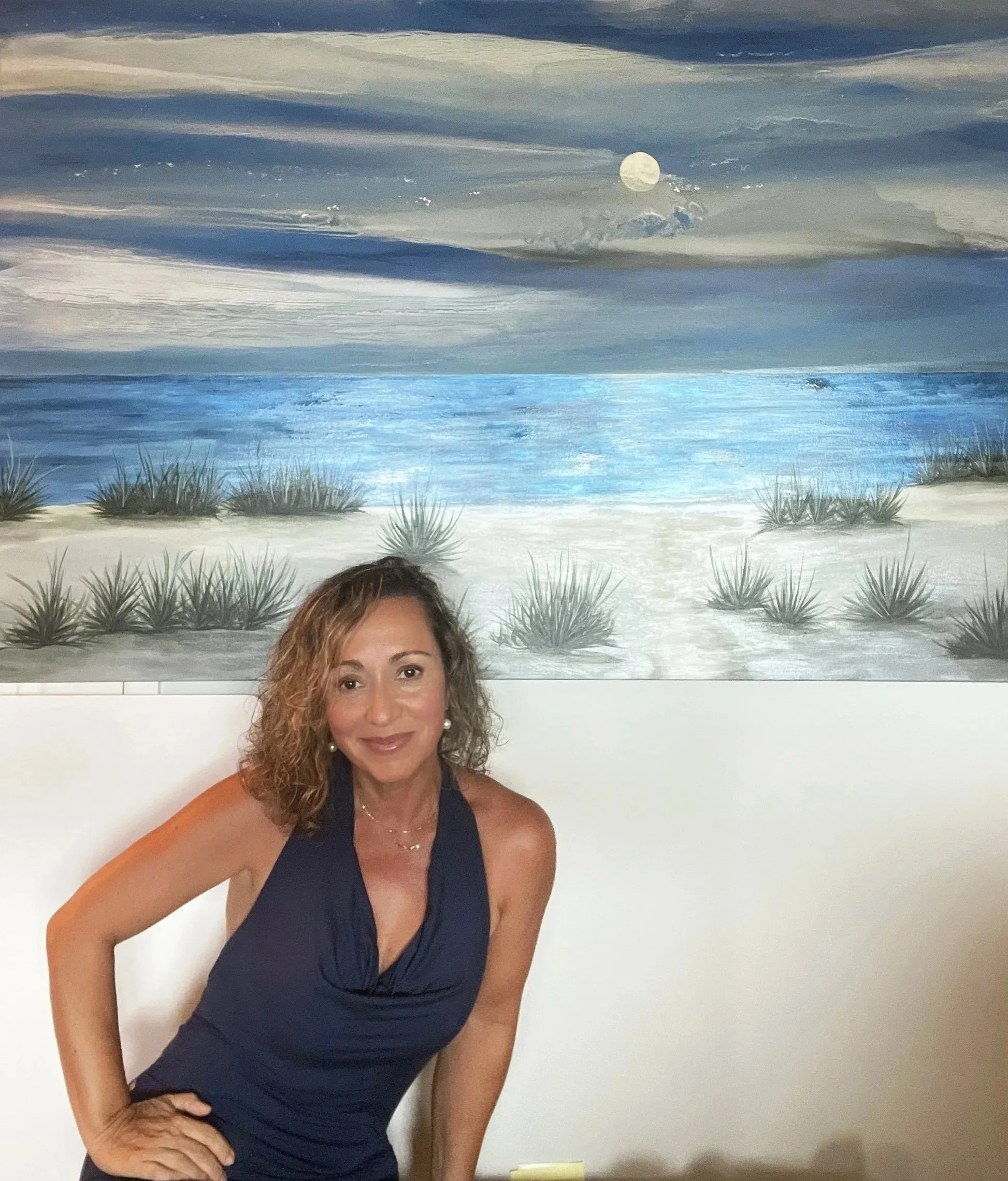 A woman with curly light brown hair wearing a navy sleeveless dress standing in front of a beach-themed painting featuring a moonlit sky, ocean, and dunes with grasses.