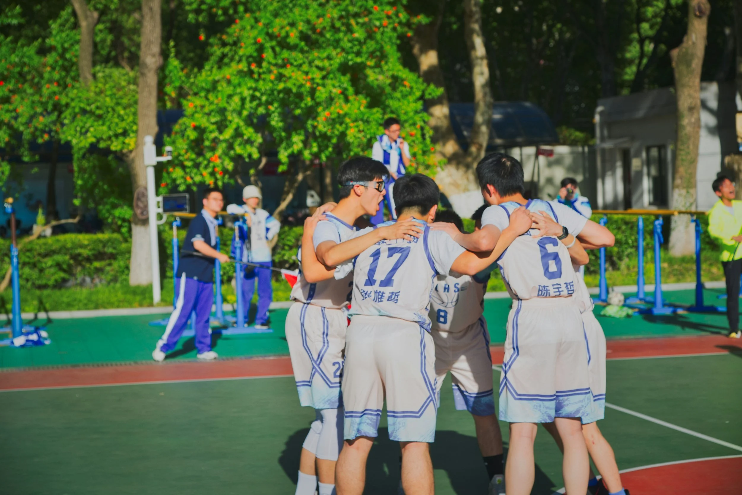 Sports Performance Anxiety Group of young basketball players huddling together on an outdoor court during daytime, with other players and a red and green court background, and trees with green foliage. Discussing prevention of sports performance anxiety