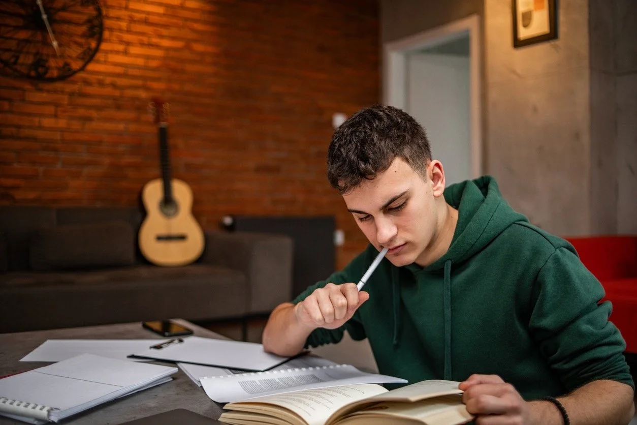 Hypnosis For Exam Anxiety Study Skills and Test Anxiety - young man in a green hoodie studying at a table with books, papers, and a pen, with an acoustic guitar hanging on a brick wall in the background.