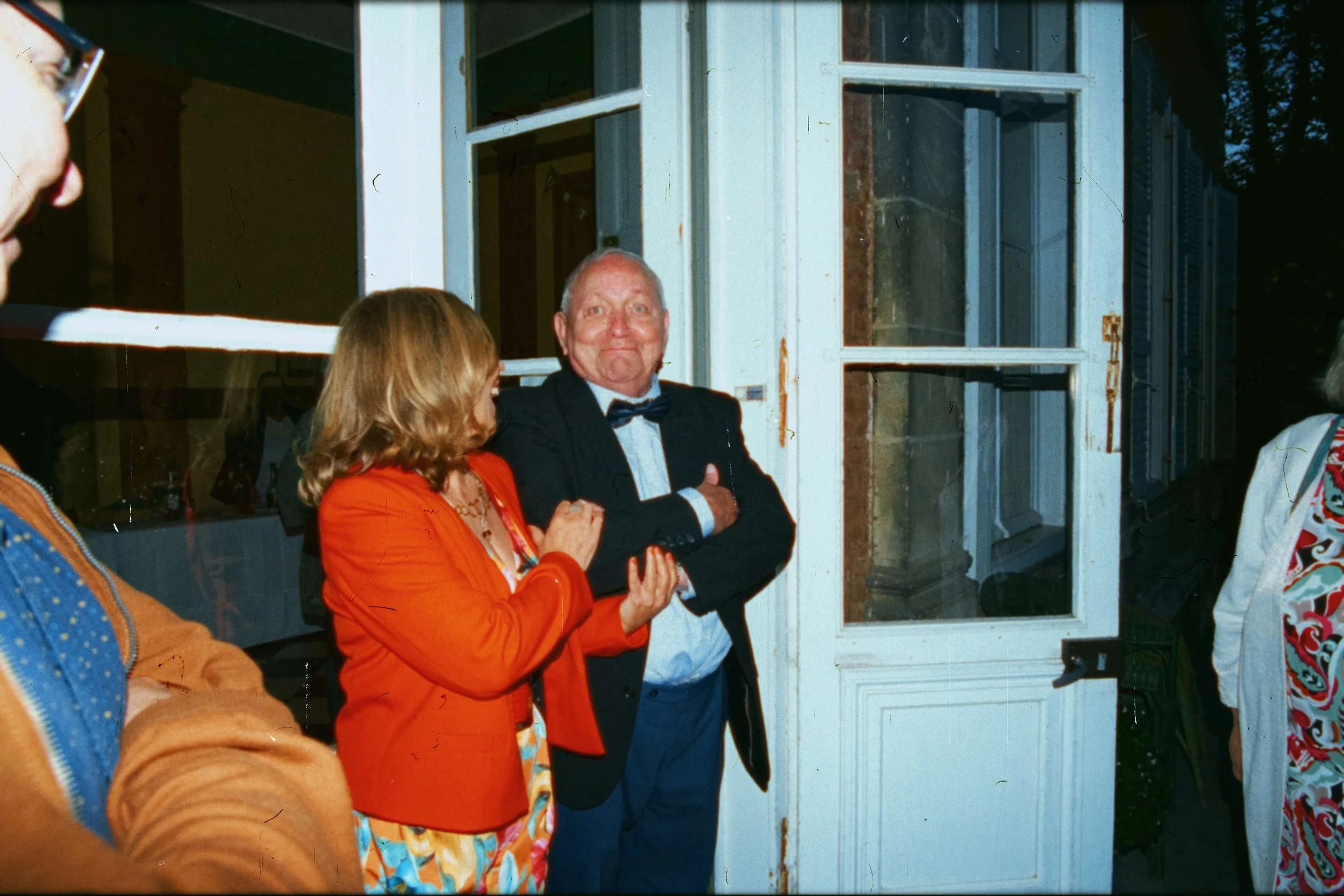 Un groupe de personnes lors d'une fête ou d'une réunion sociale, avec un homme en costume noir avec un nœud papillon et une femme en robe colorée et veste rouge, près d'une porte en bois blanc avec plusieurs fenêtres.
