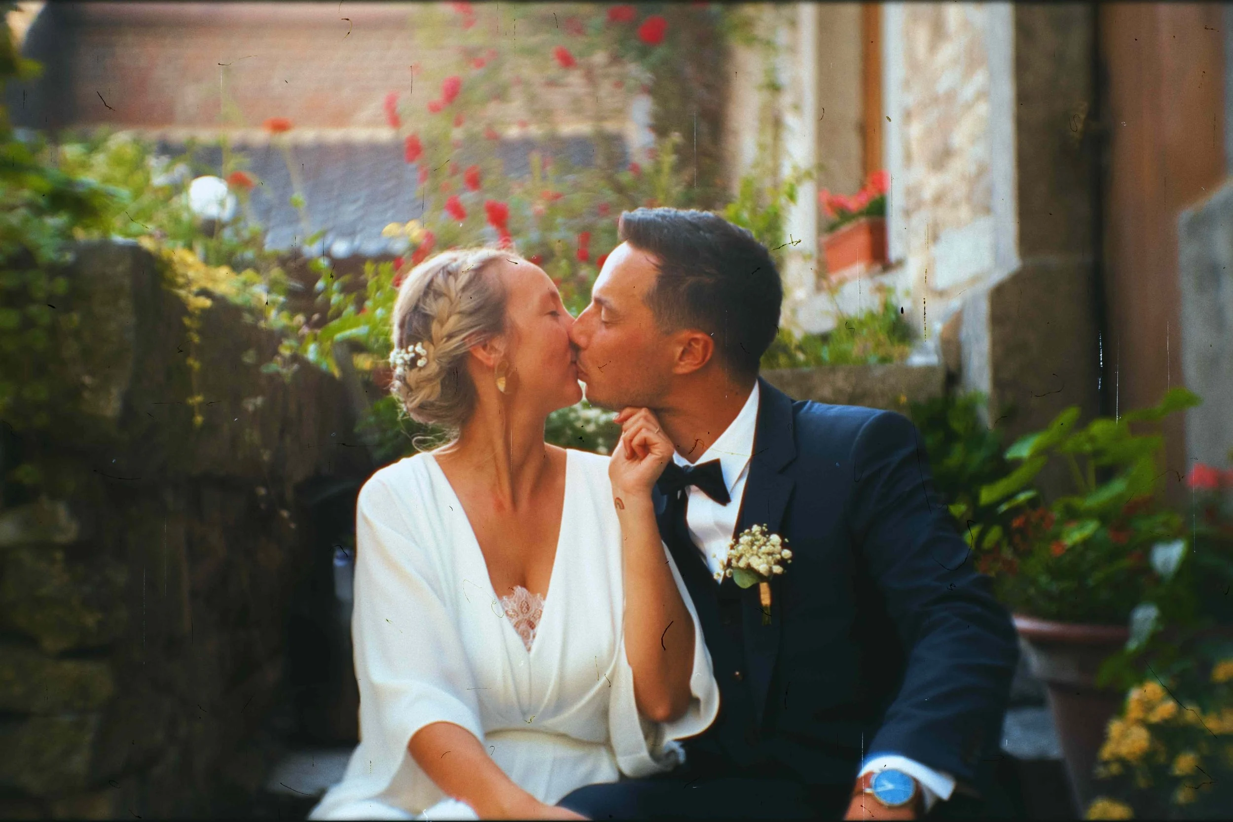 Un couple en costume de mariage, une femme en robe blanche et un homme en costume noir avec nœud papillon, échangeant un baiser dans un jardin fleuri.