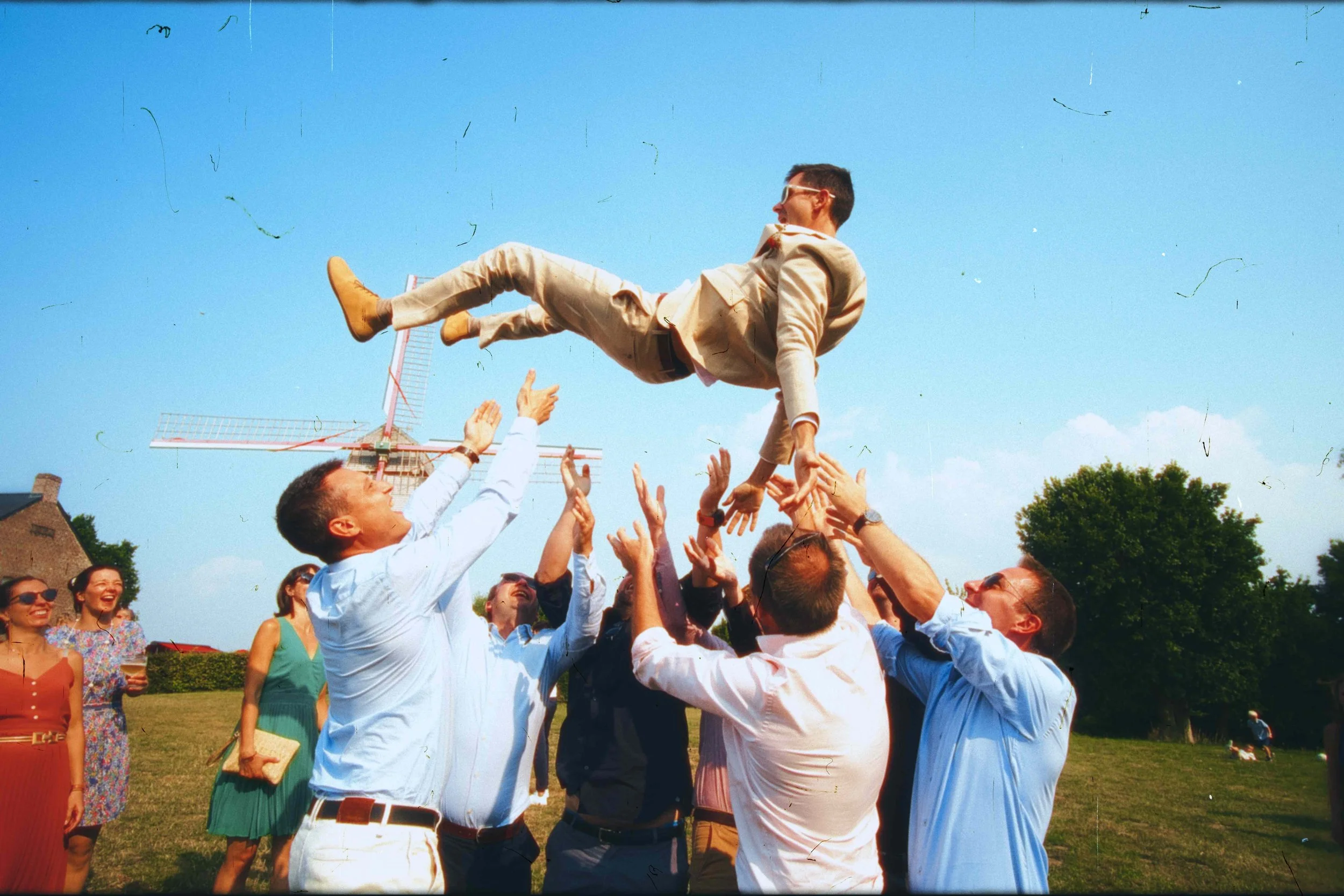 Groupe de personnes en extérieur, celebrant lors d'une fête ou d'un mariage, en train de lancer un homme dans les airs contre un ciel bleu clair.