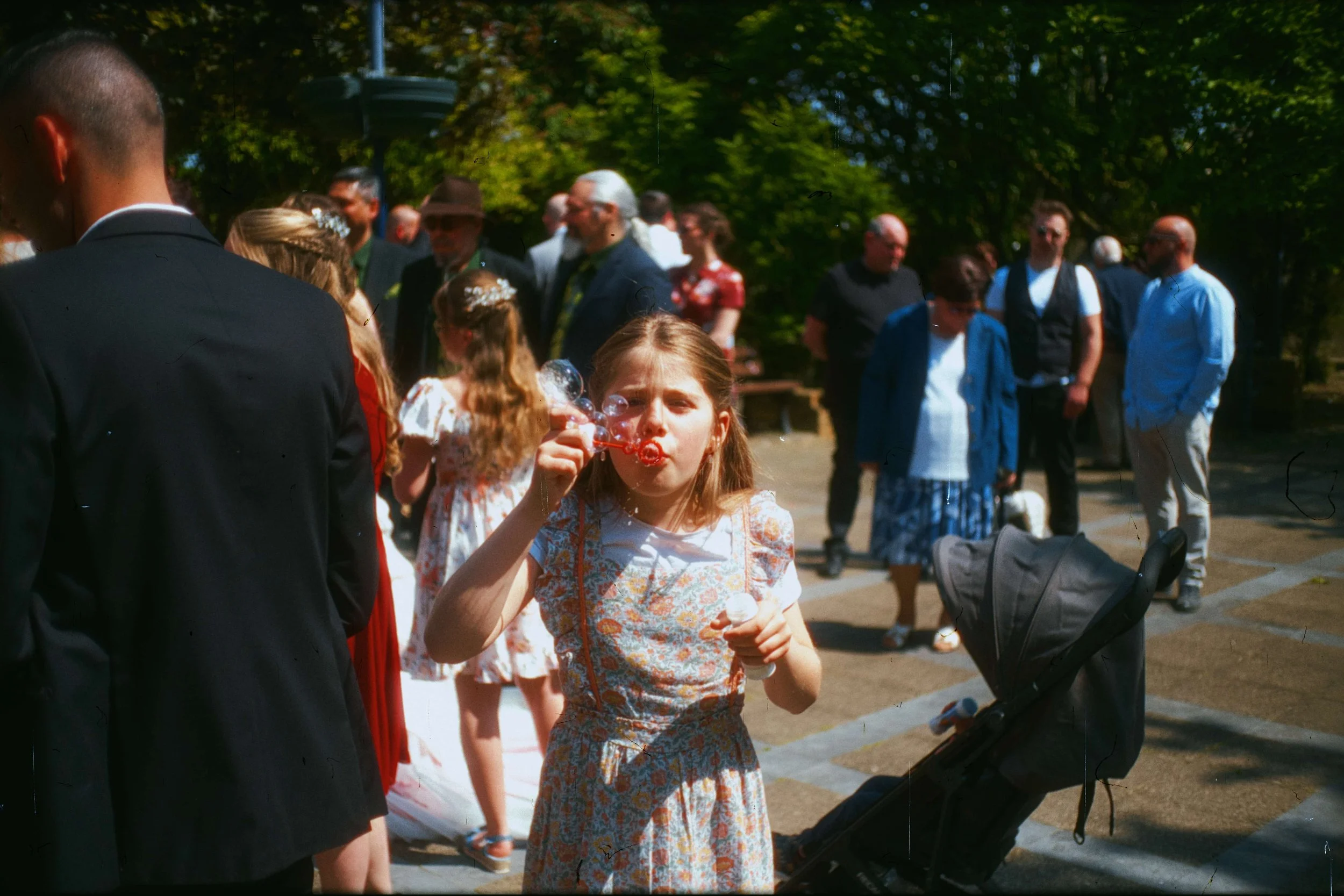 Une jeune fille souffle des bulles lors d'un rassemblement en plein air, entourée de plusieurs adultes et enfants, avec des arbres en fond.