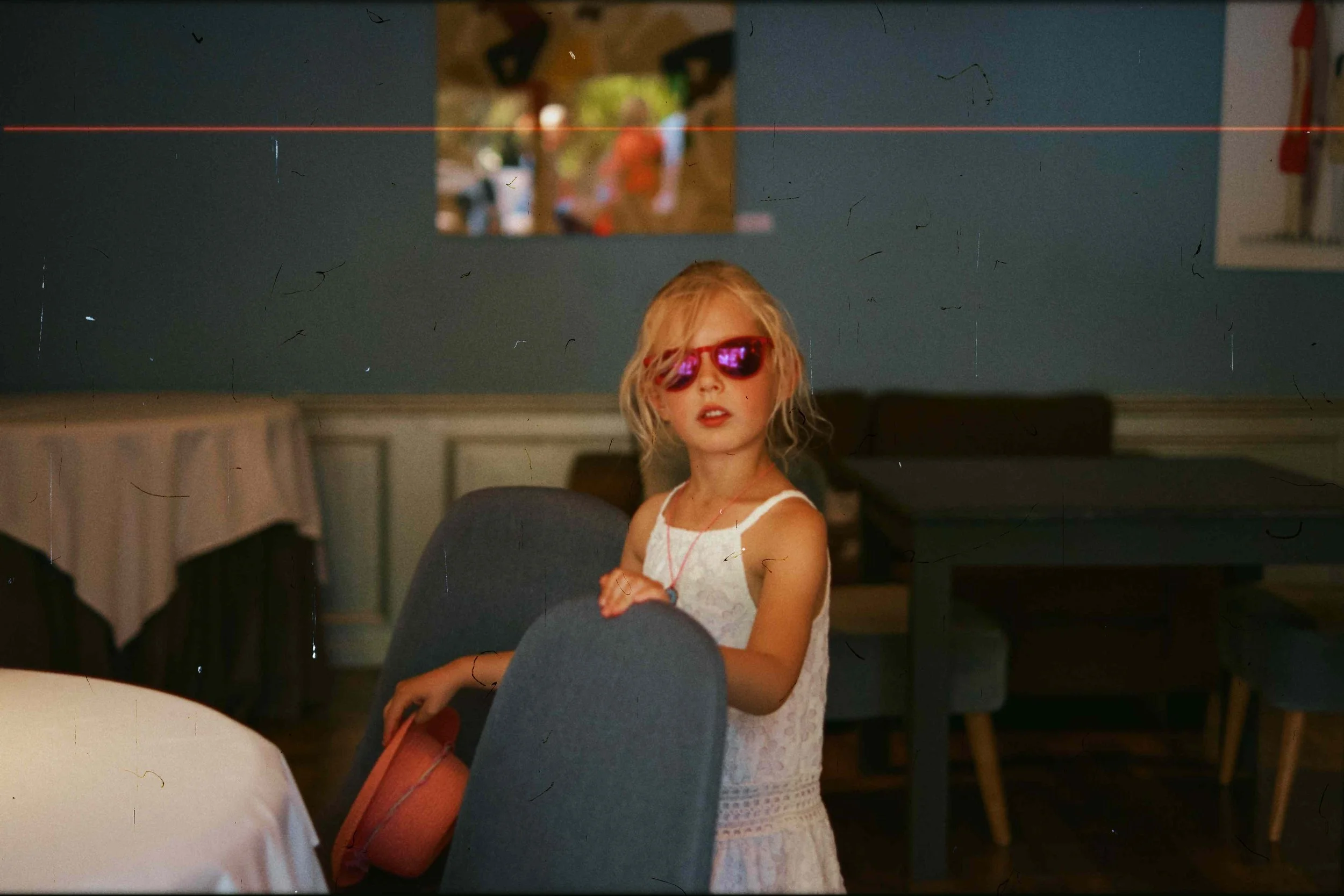 Jeune fille avec des lunettes de soleil roses, portant une robe blanche, dans un restaurant ou une salle de repas, tenant une chaise