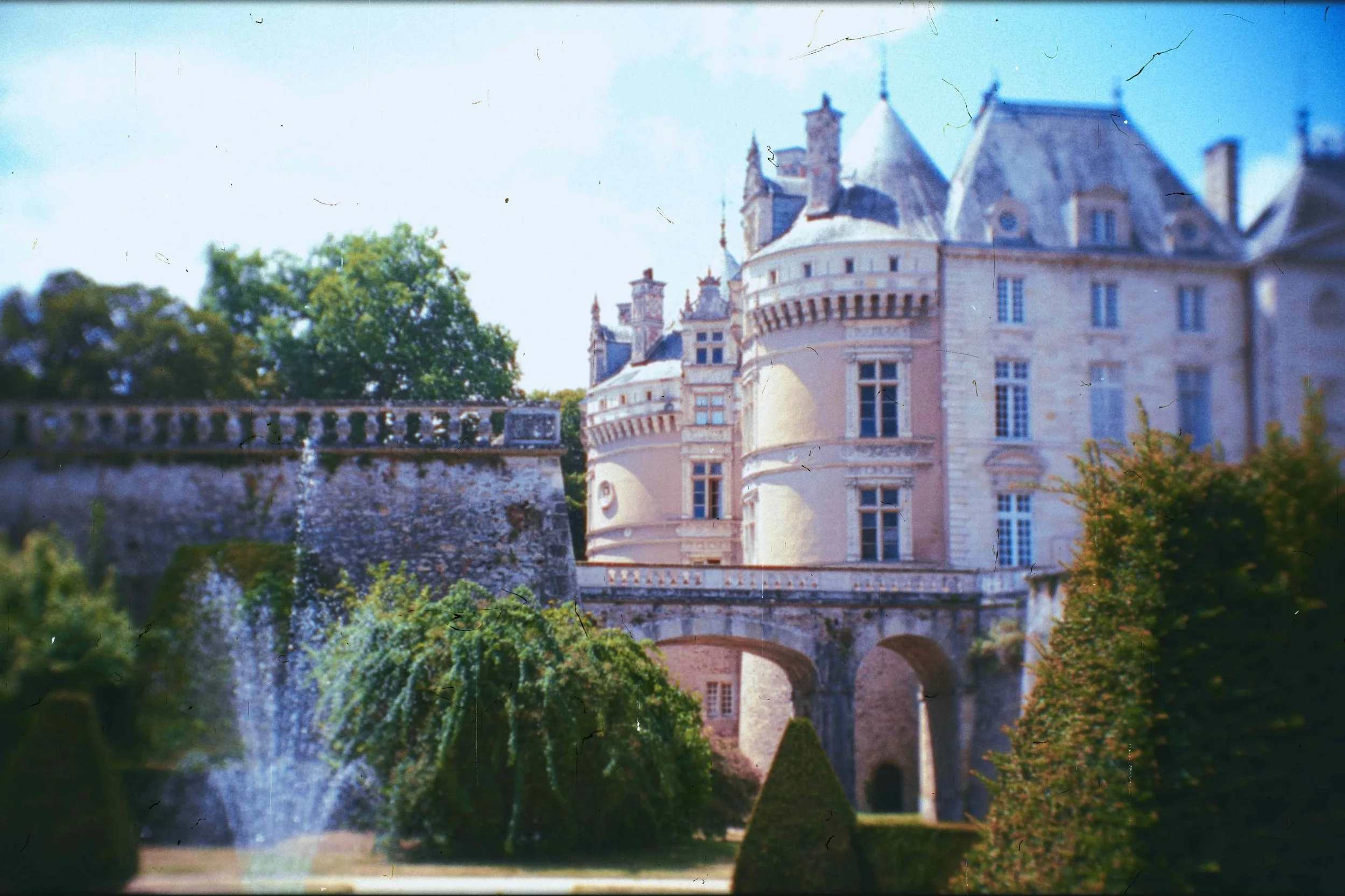 Château historique avec tours et tourelles, entouré de jardins verdoyants et d'un bâtiment avec un pont en arc sous lequel passe un petit ruisselet, sous un ciel bleu ensoleillé.