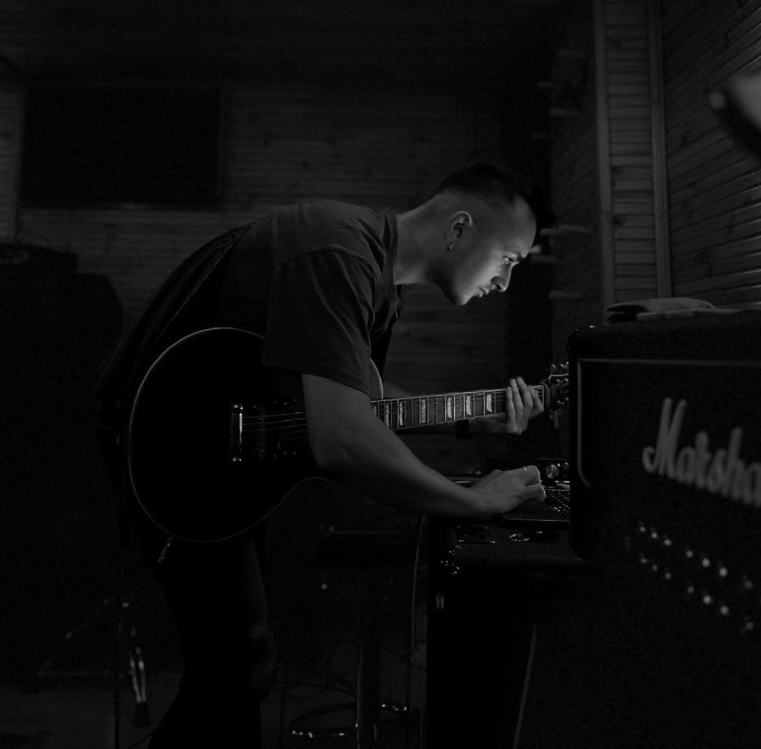 A young man playing an acoustic guitar while working on a keyboard or electronic equipment in a dimly lit music studio.