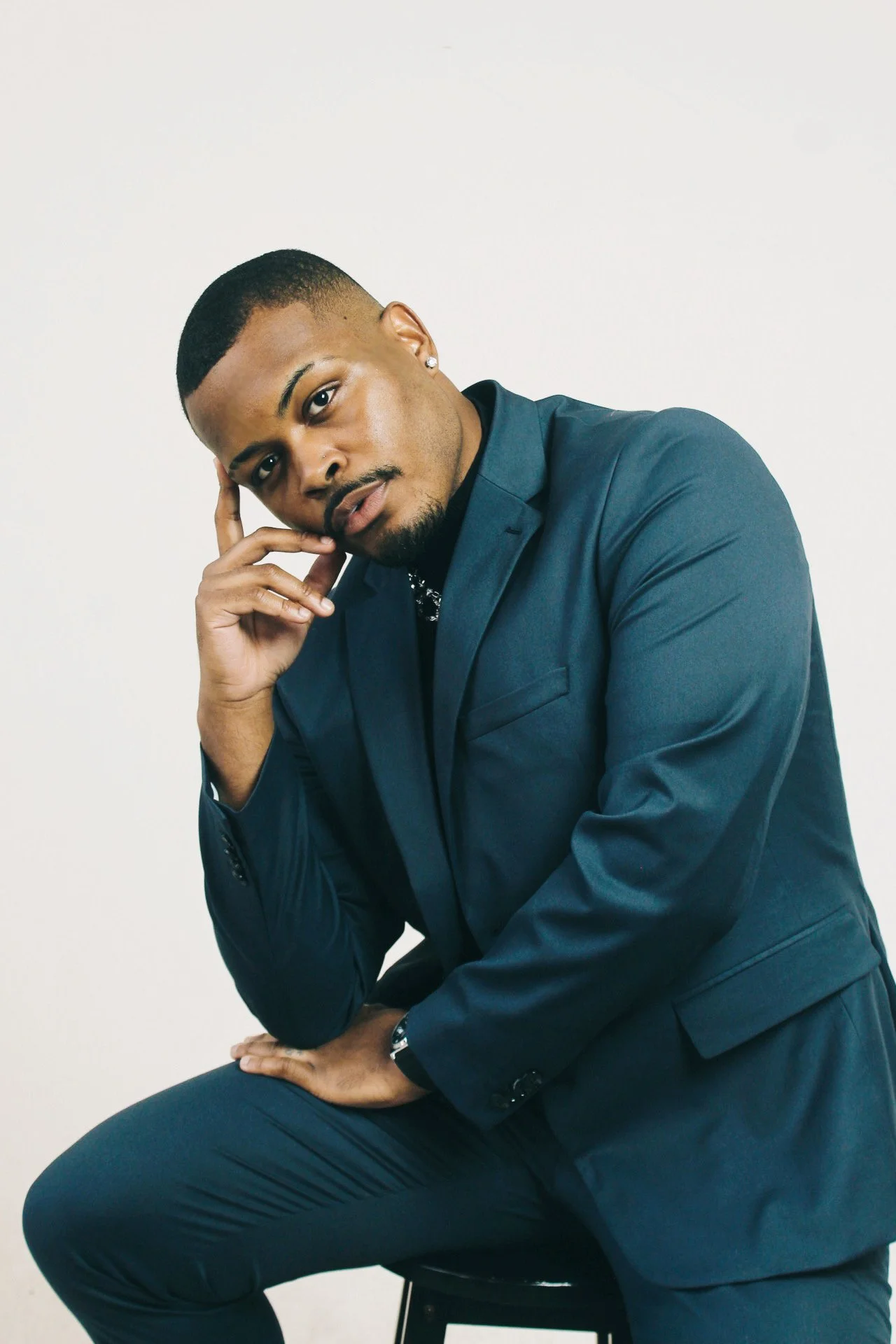 A young man with a short haircut and earrings, wearing a dark blue suit, sitting on a stool against a plain white background. He is resting his chin on his hand and looking at the camera with a thoughtful expression.