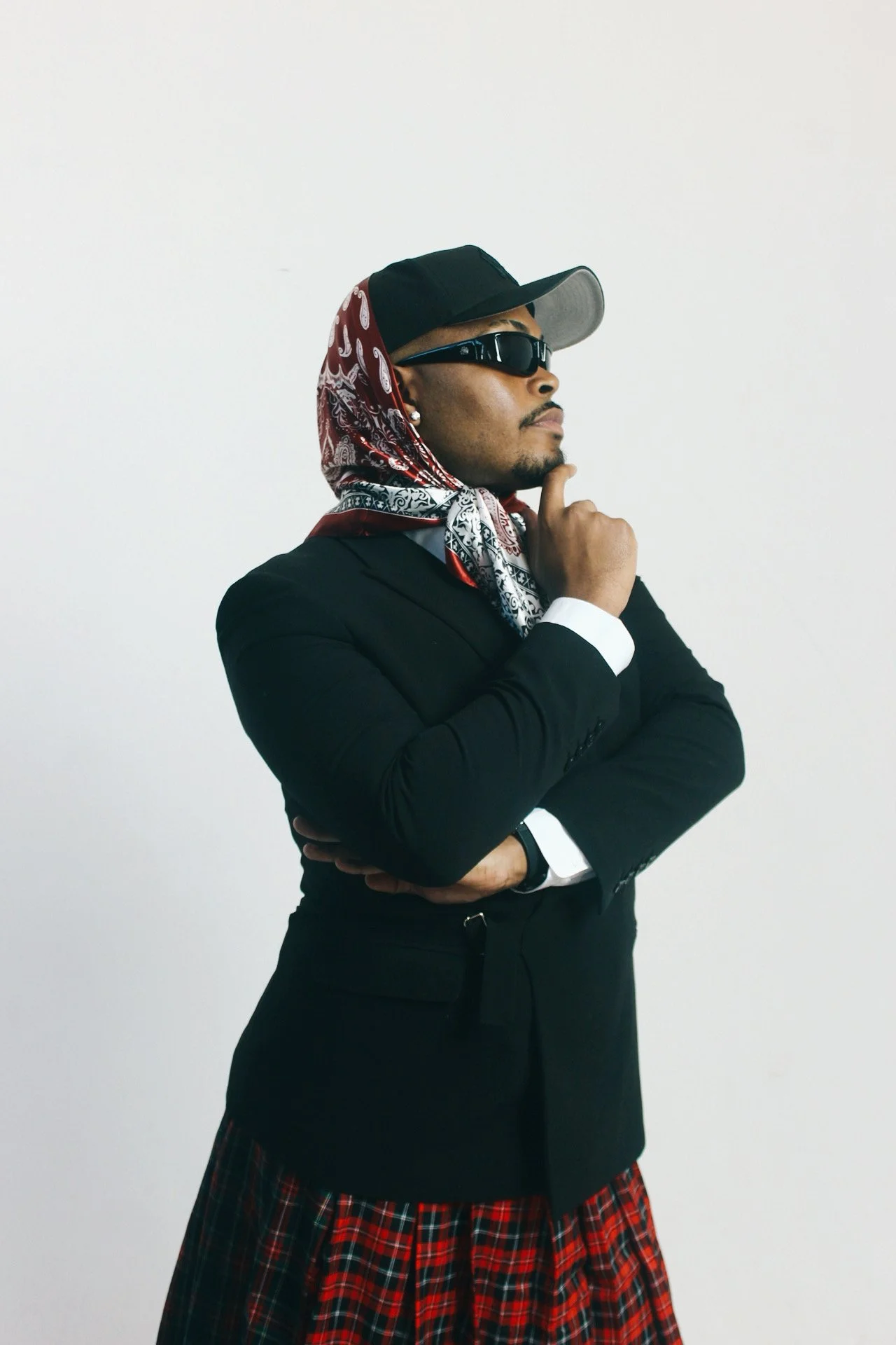 A person dressed in a black blazer and a red tartan skirt, wearing sunglasses, a cap, a bandana, and posing with a thoughtful expression against a plain white background.