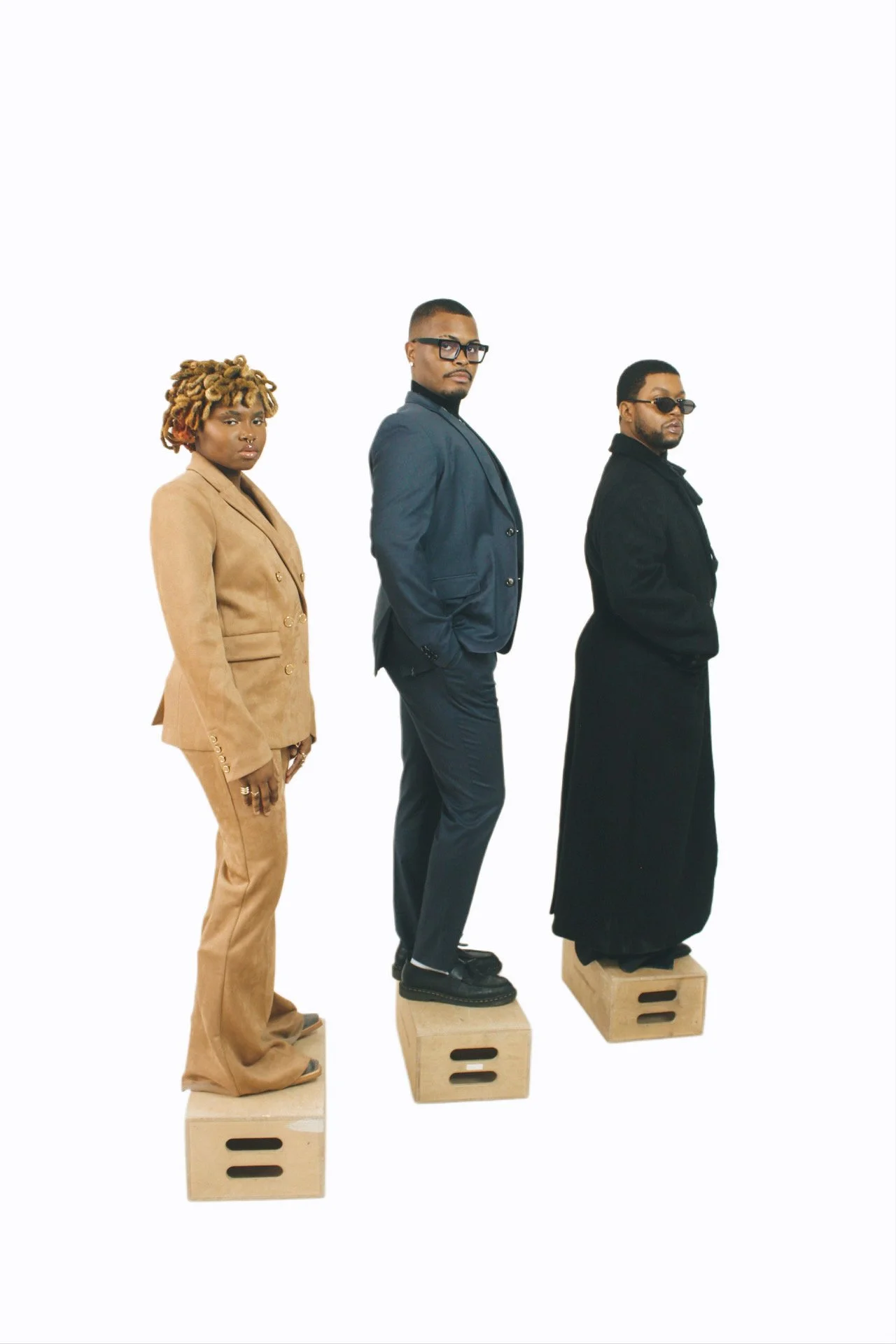 Three individuals standing on wooden blocks in a line, dressed in stylish outfits, with a plain white background.