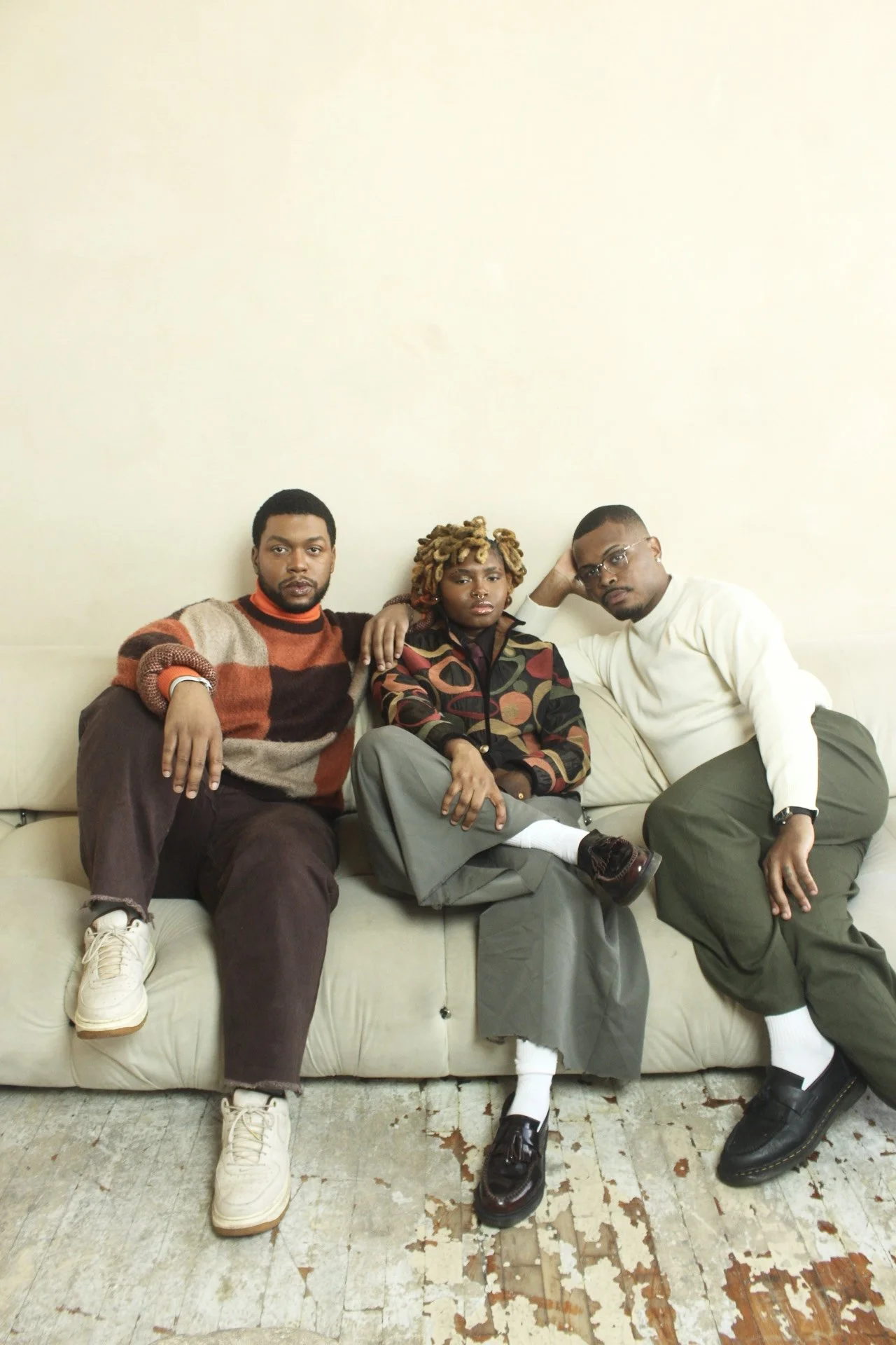 Three people sitting on a cream sofa in a room with a plain wall, dressed casually in vintage-style clothing.