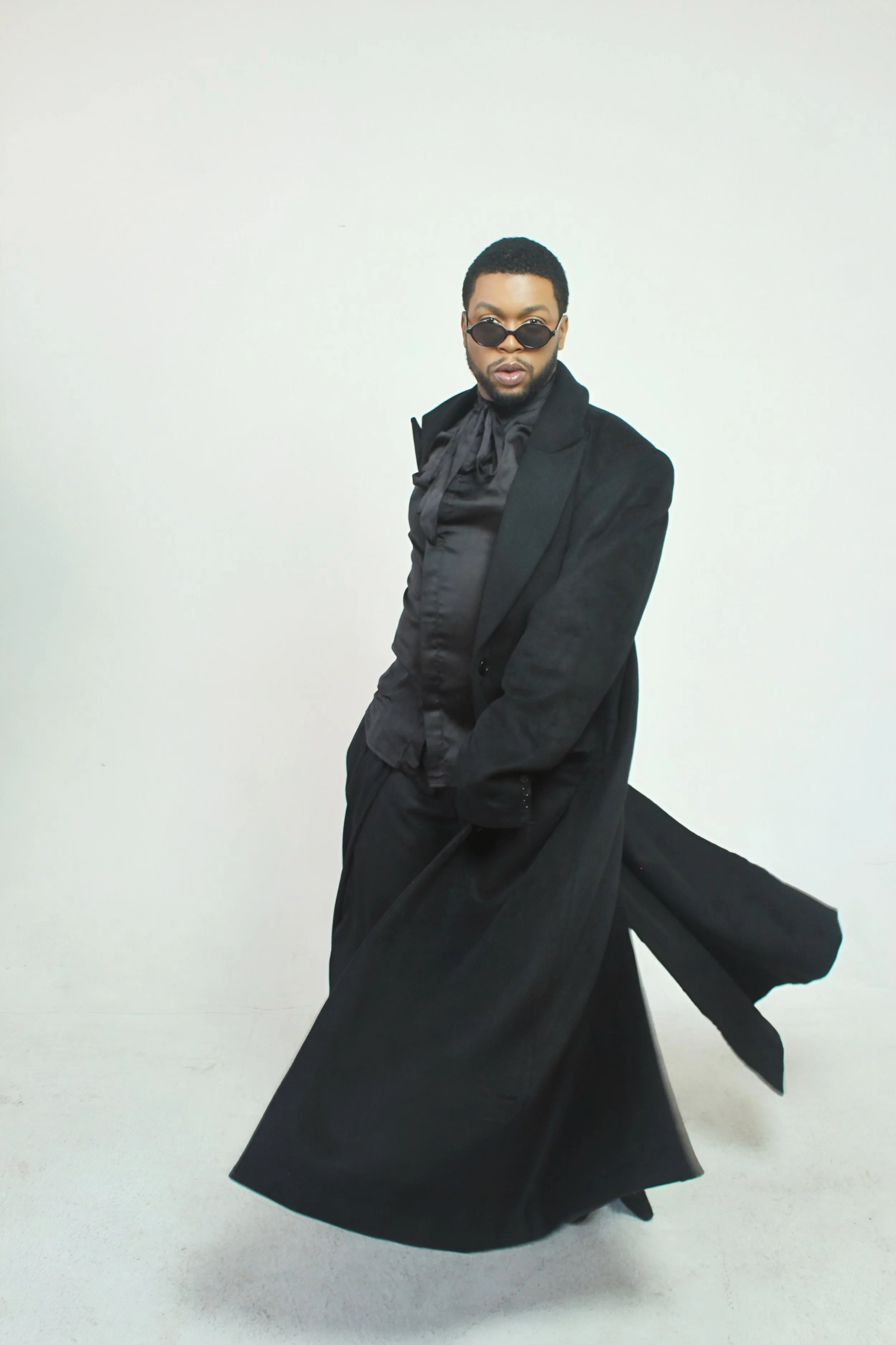 A man dressed in a long black coat and sunglasses, walking confidently against a white background.