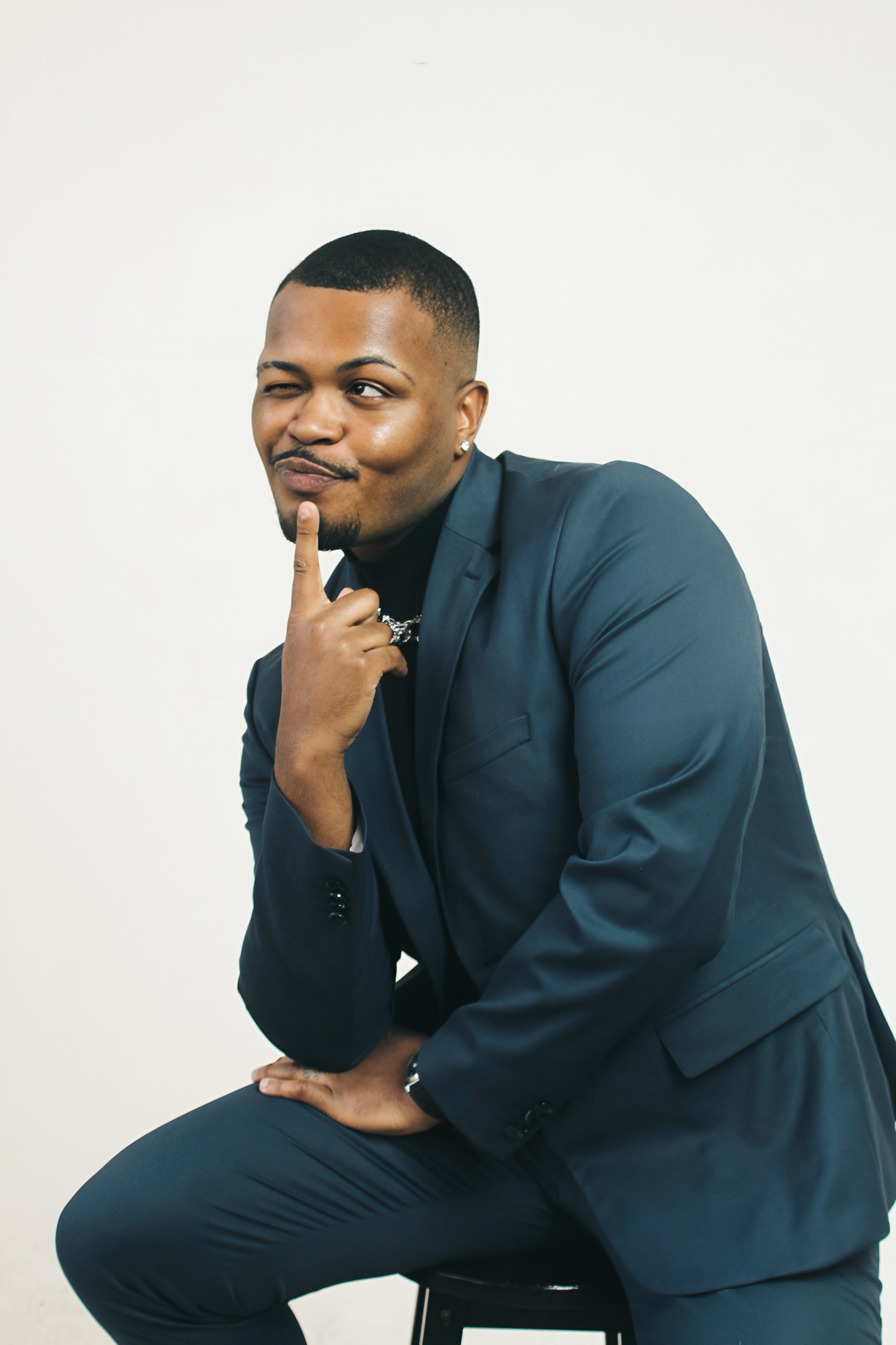 A man in a dark blue suit sitting on a stool, with one hand raised near his face and his index finger touching his chin, looking playful with a slight smile and a wink.