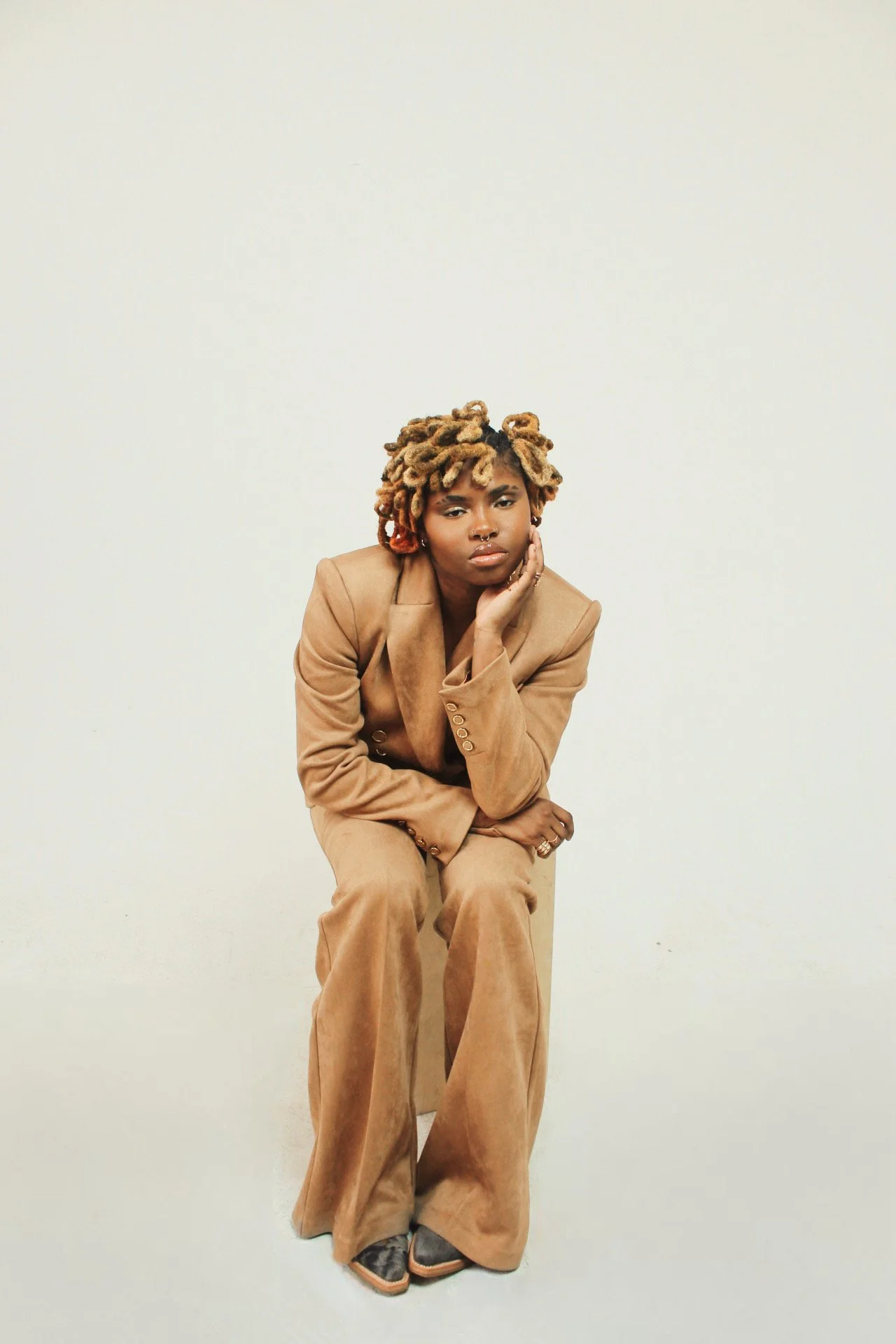 A woman in a tan suit sits on a small cube against a plain white background, resting her chin on her hand and looking thoughtfully at the camera.