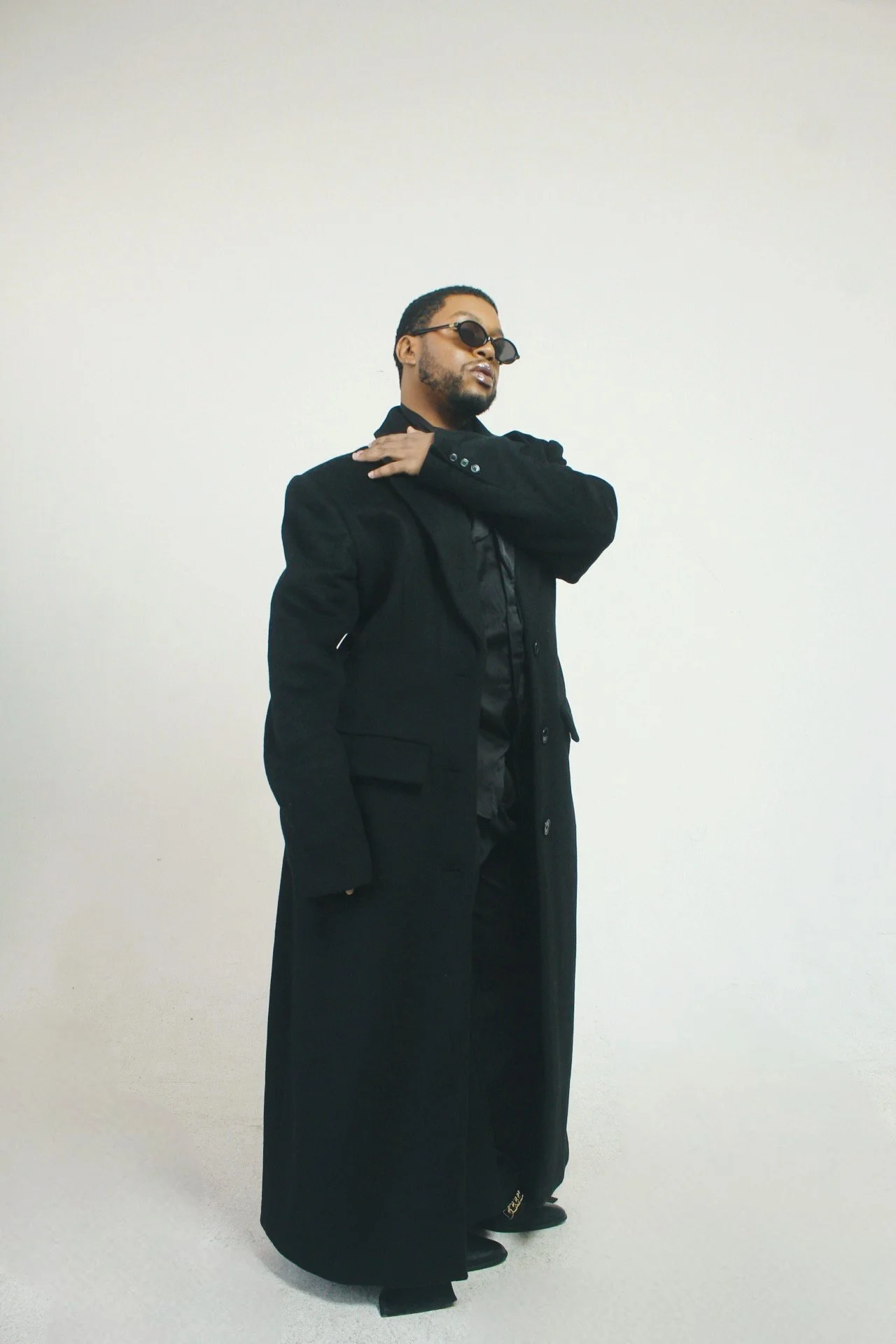 A man wearing black sunglasses, a long black coat, and black clothing, standing against a plain white wall, striking a pose with one hand on his shoulder.