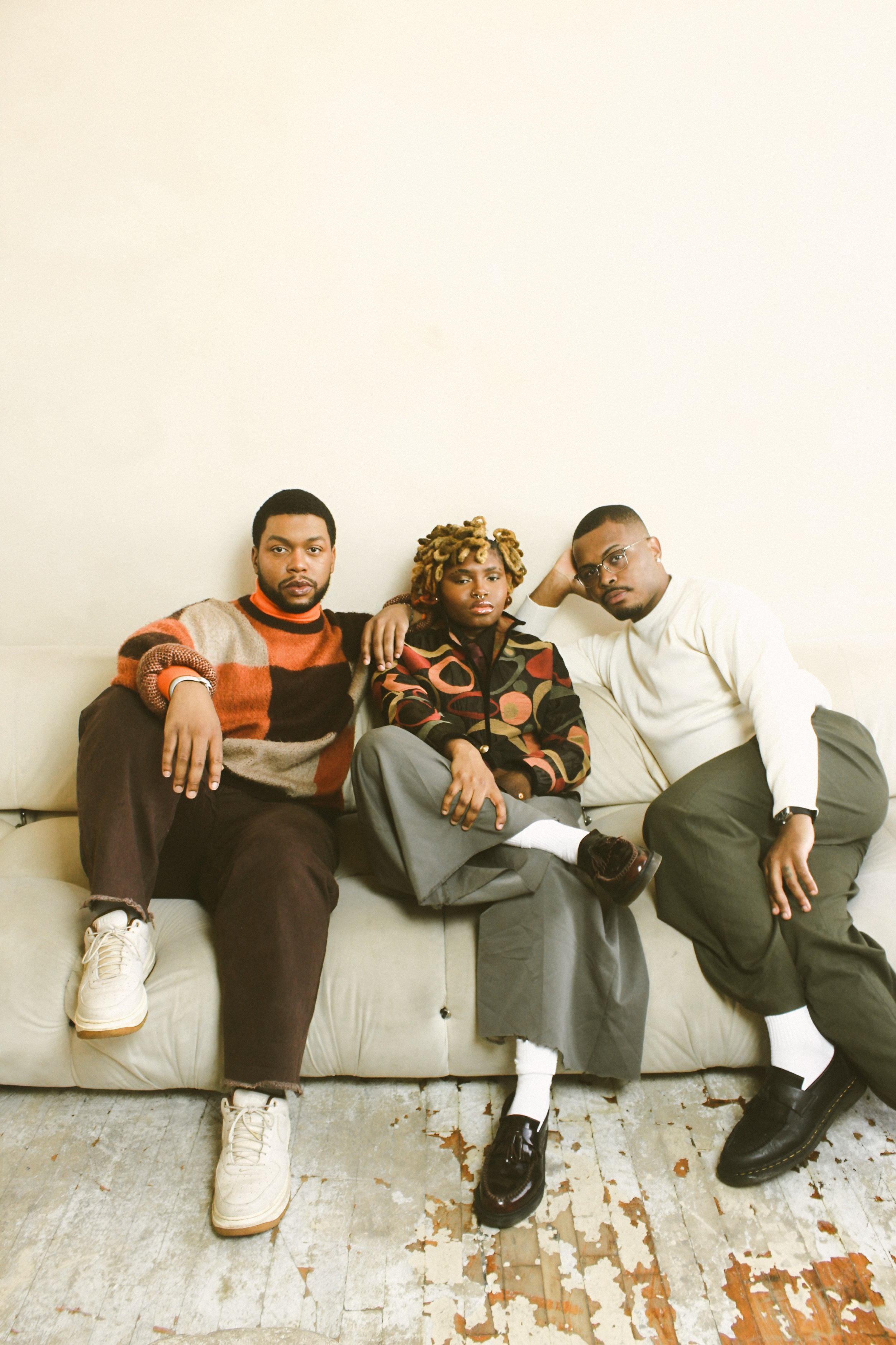 Three people sitting on a beige couch against a white wall, with a worn wooden floor visible.