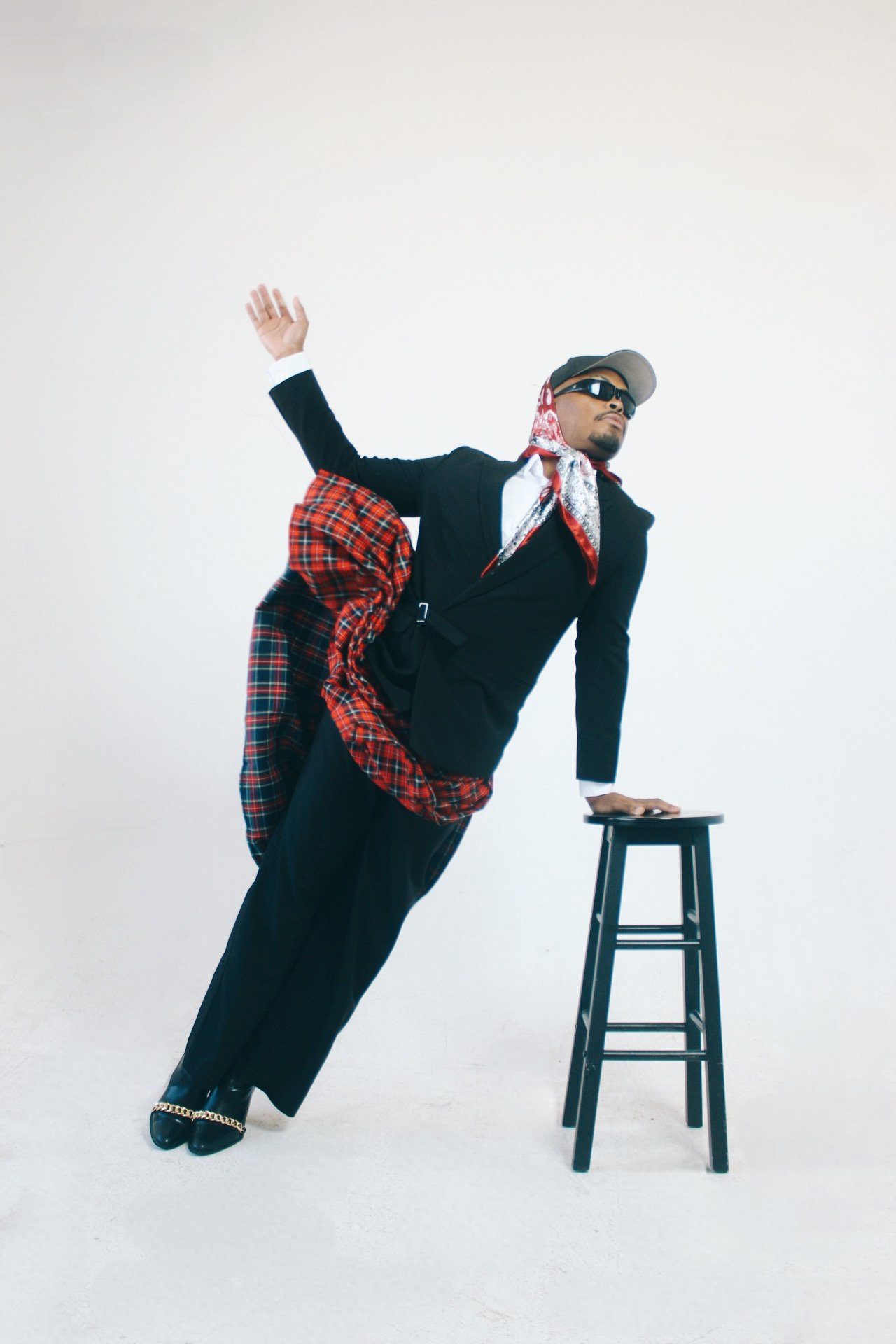 A person in a black suit with extra layers of clothing, such as a red plaid skirt, standing with one hand on a stool and the other raised, wearing sunglasses and a hat against a plain white background.